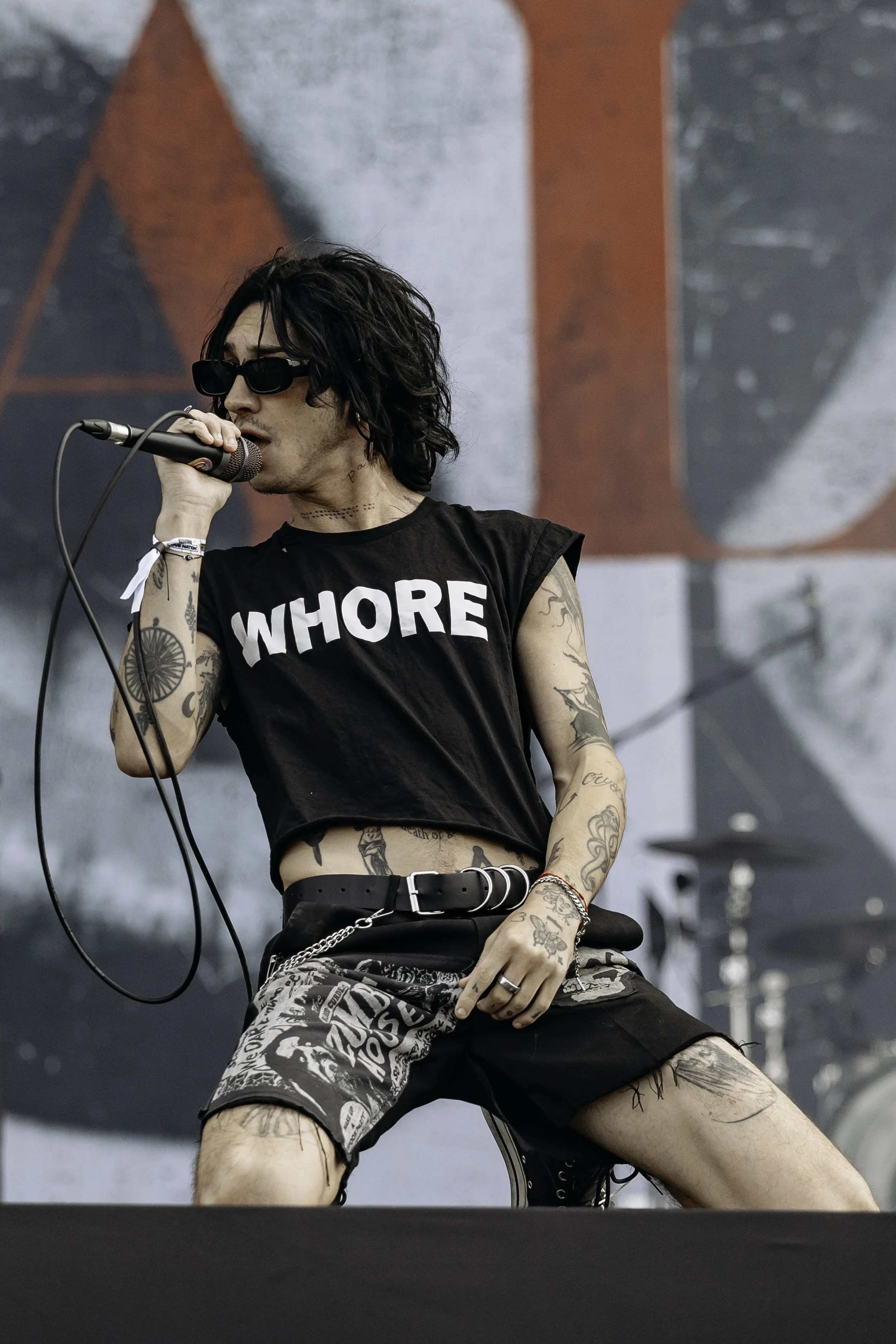 A musician with black curly hair, sunglasses, tattoos, and wearing a black sleeveless shirt with the word 'WHORE' sings into a microphone during a performance on an outdoor stage.