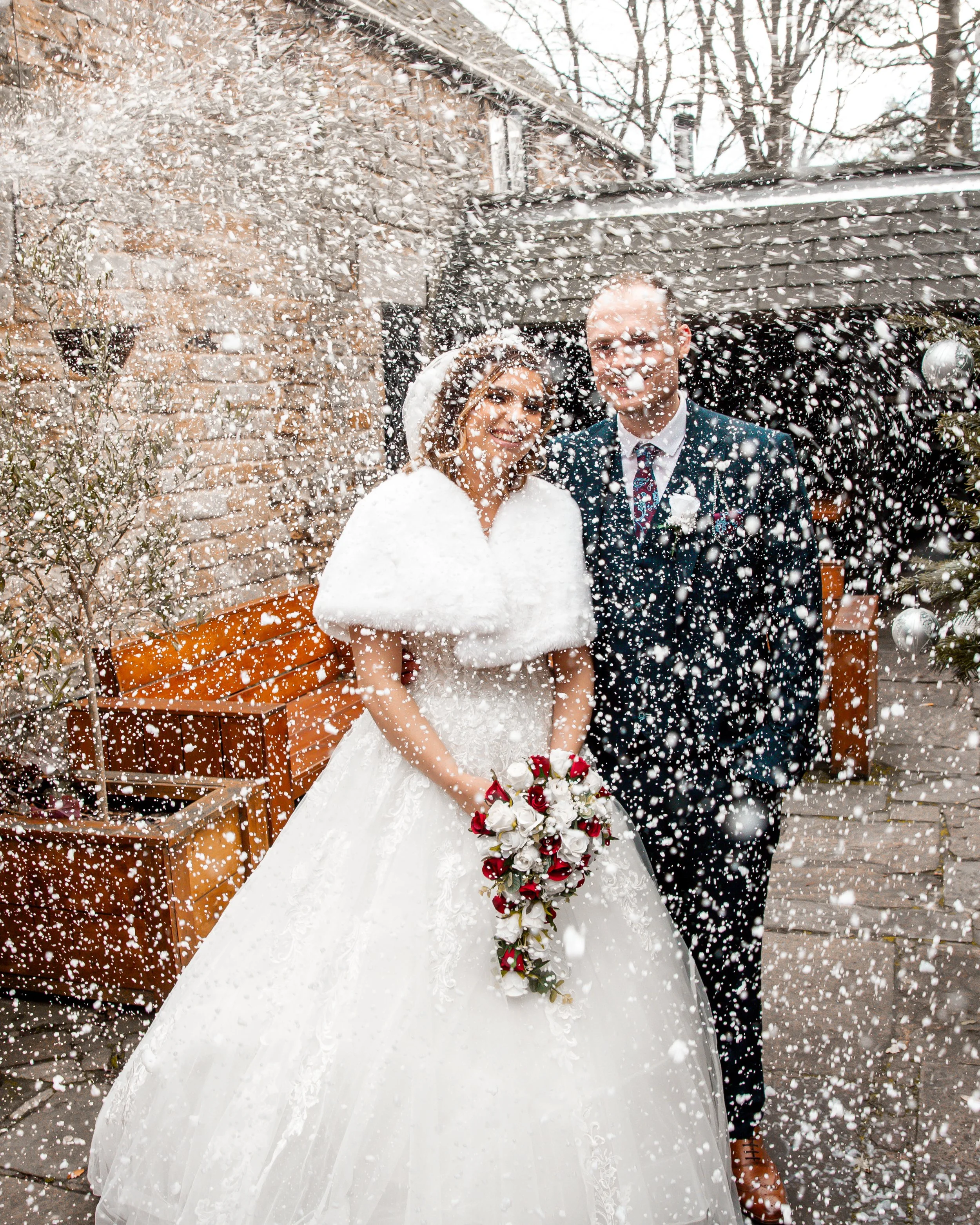 Bride and groom celebrating wedding outdoors with snow falling, bride holding a bouquet of red and white flowers, bride in white gown with cape, groom in dark suit with patterned tie.