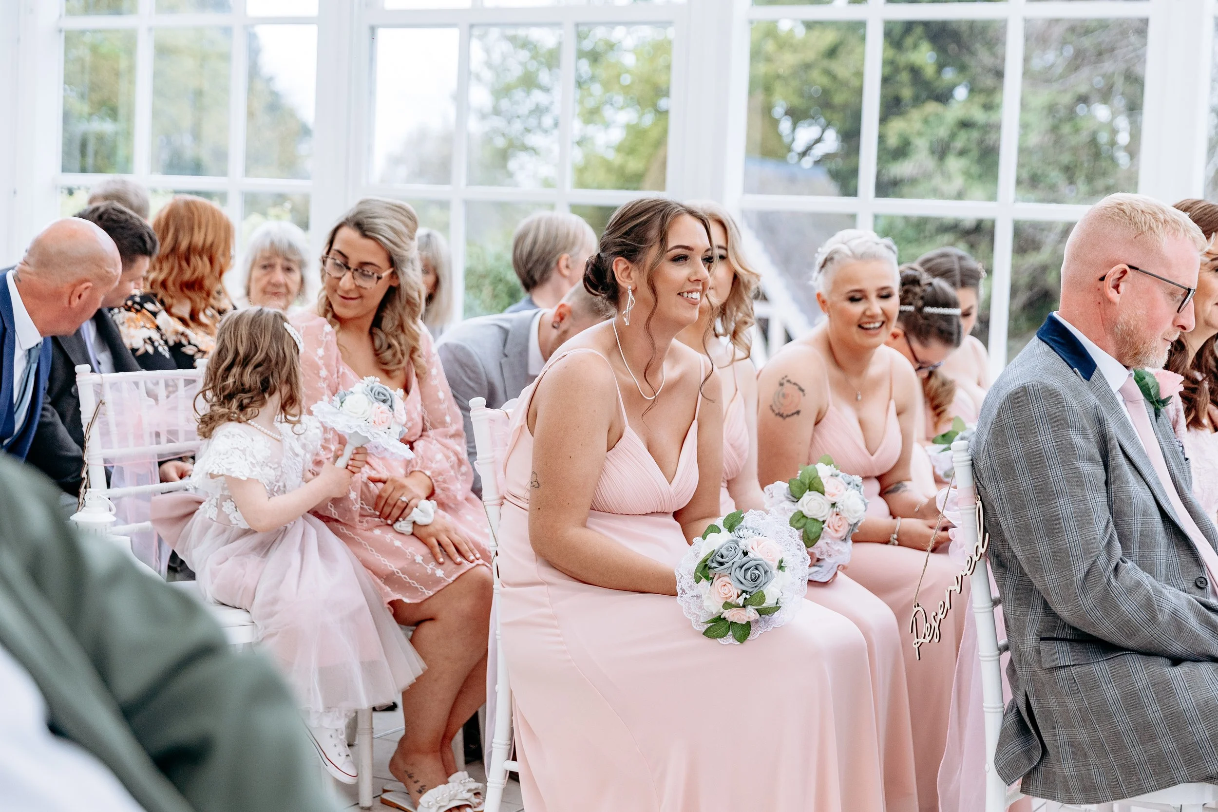 People attending a wedding ceremony seated indoors with large windows in the background, some holding bouquets, dressed in pastel and formal attire.