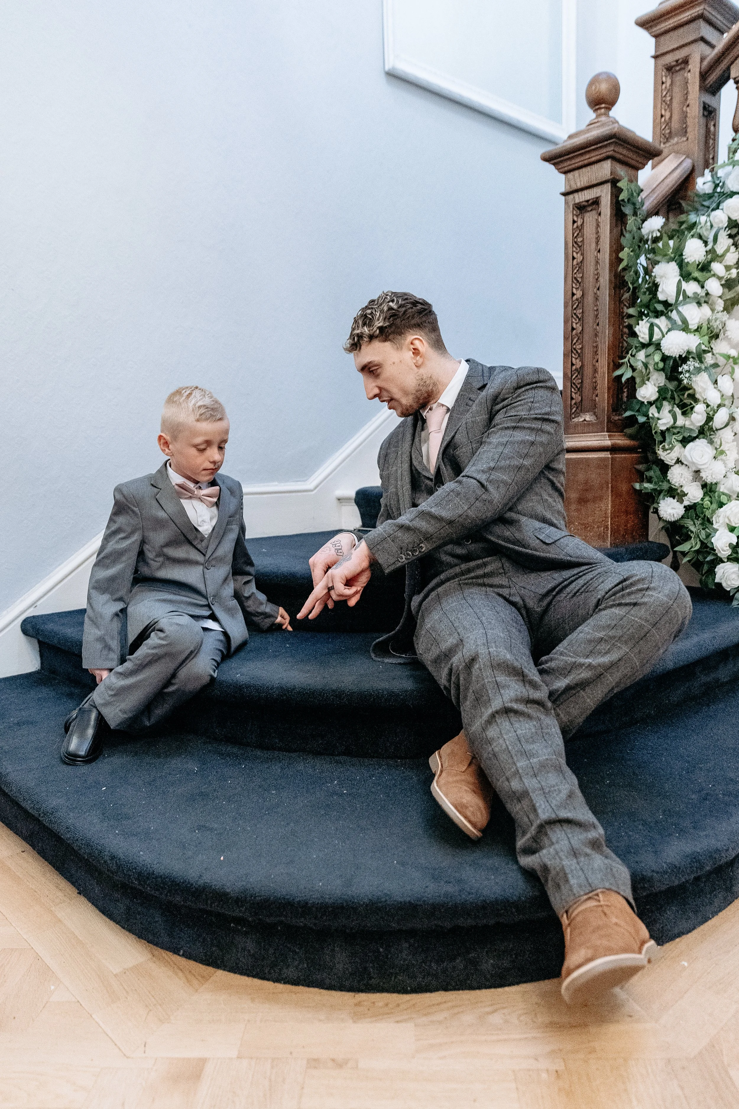 A man and young boy in gray suits sitting on a staircase, having a conversation. The man is pointing at something with his finger, and both are dressed formally for a special occasion.
