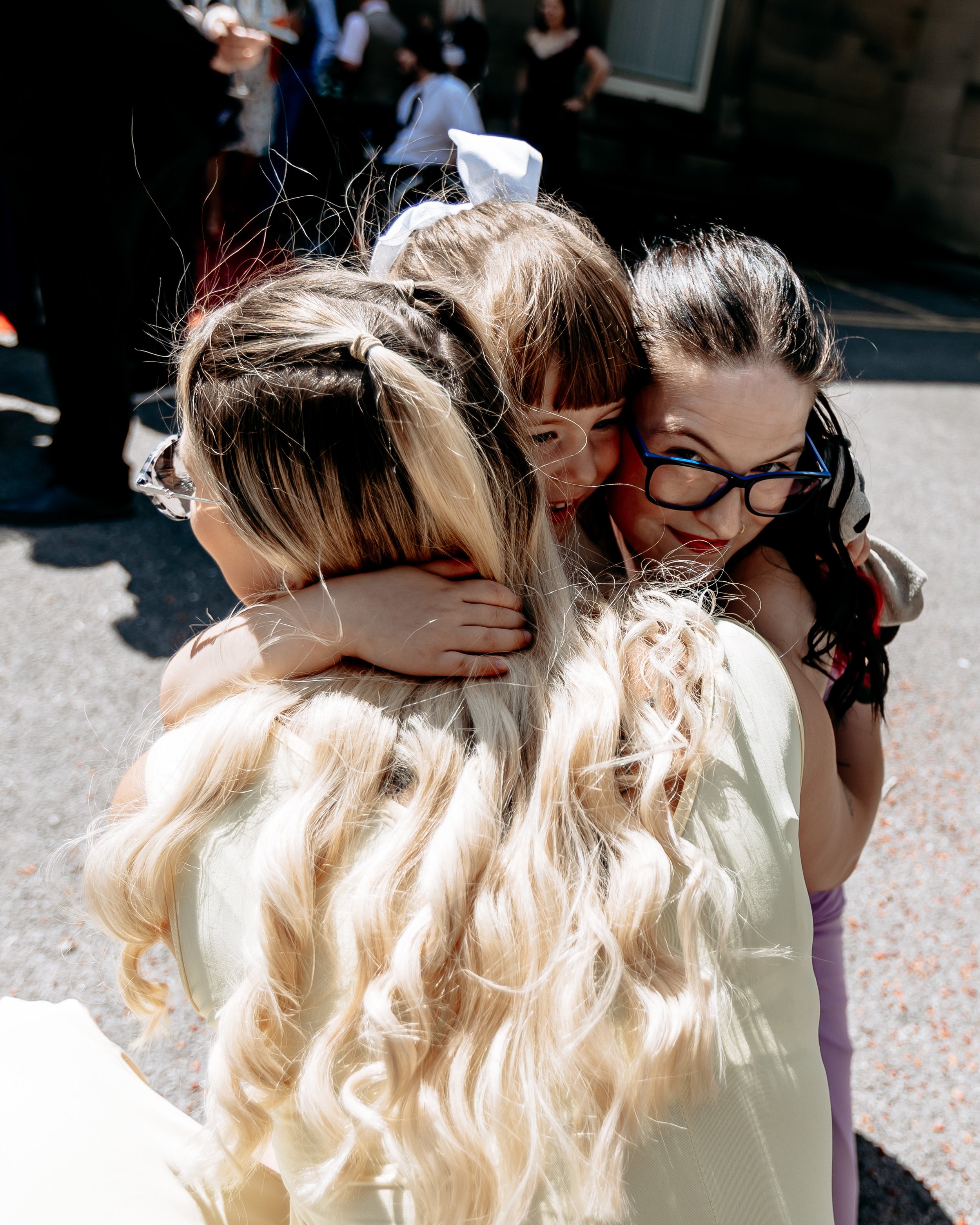 A group of women and girls hugging outdoors on a sunny day, with some wearing glasses and others with long, wavy blonde hair.