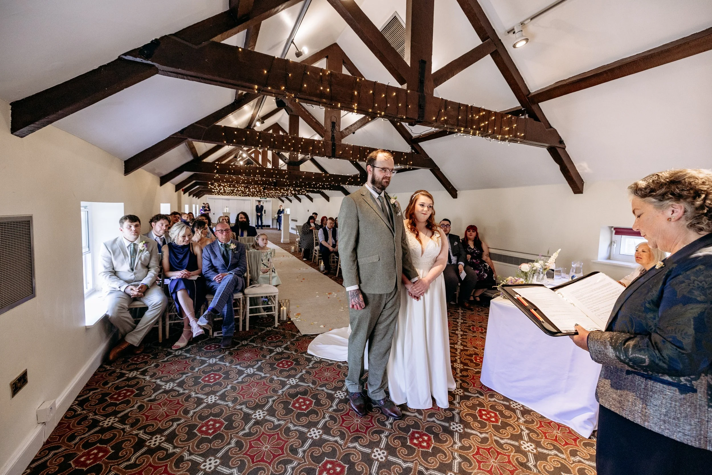 A couple stands hand-in-hand during a wedding ceremony in a decorated indoor chapel with guests seated on either side and string lights on wooden beams overhead.