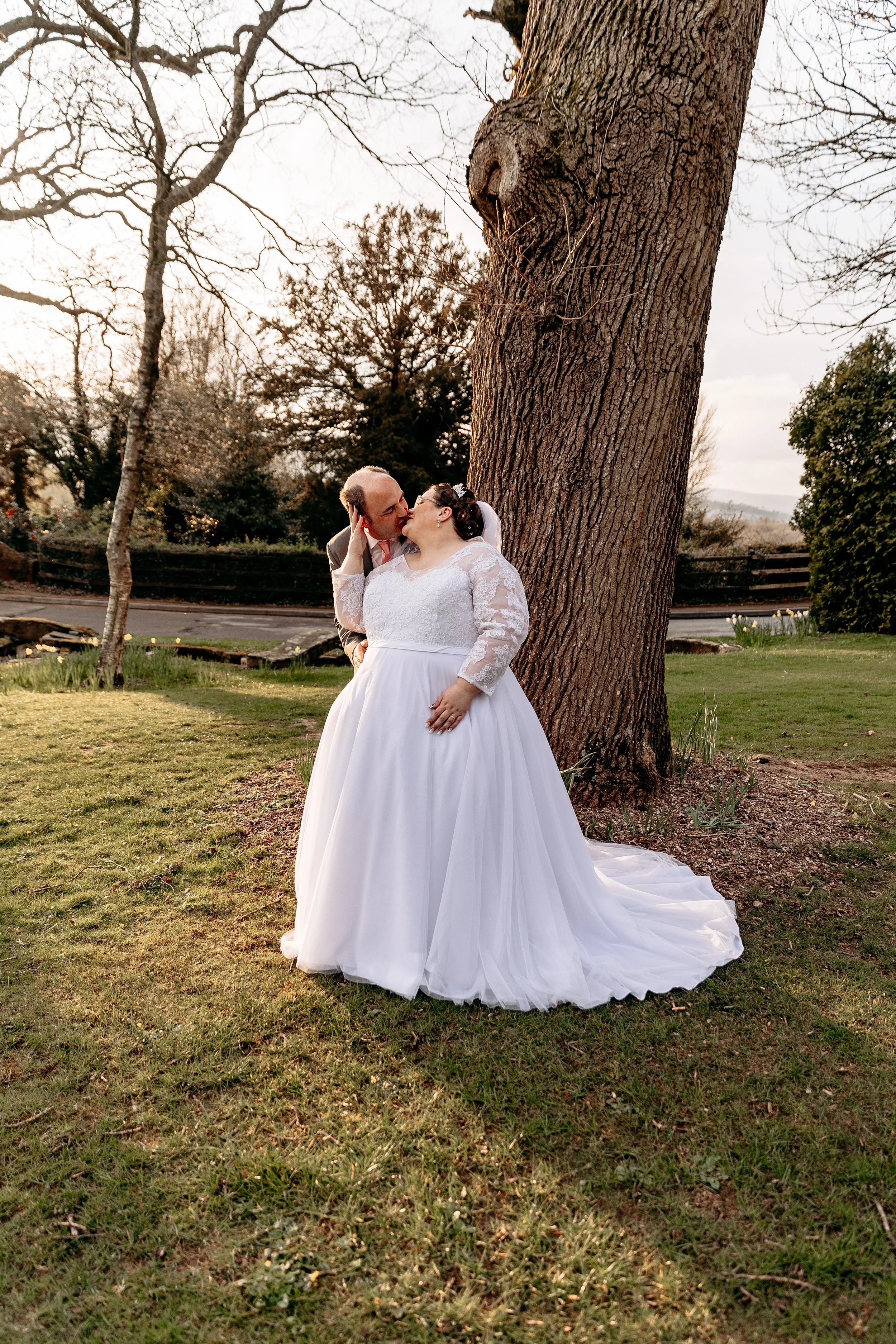A newlywed couple sharing a kiss outdoors near a large tree, with the bride in a white wedding gown and the groom in a suit.