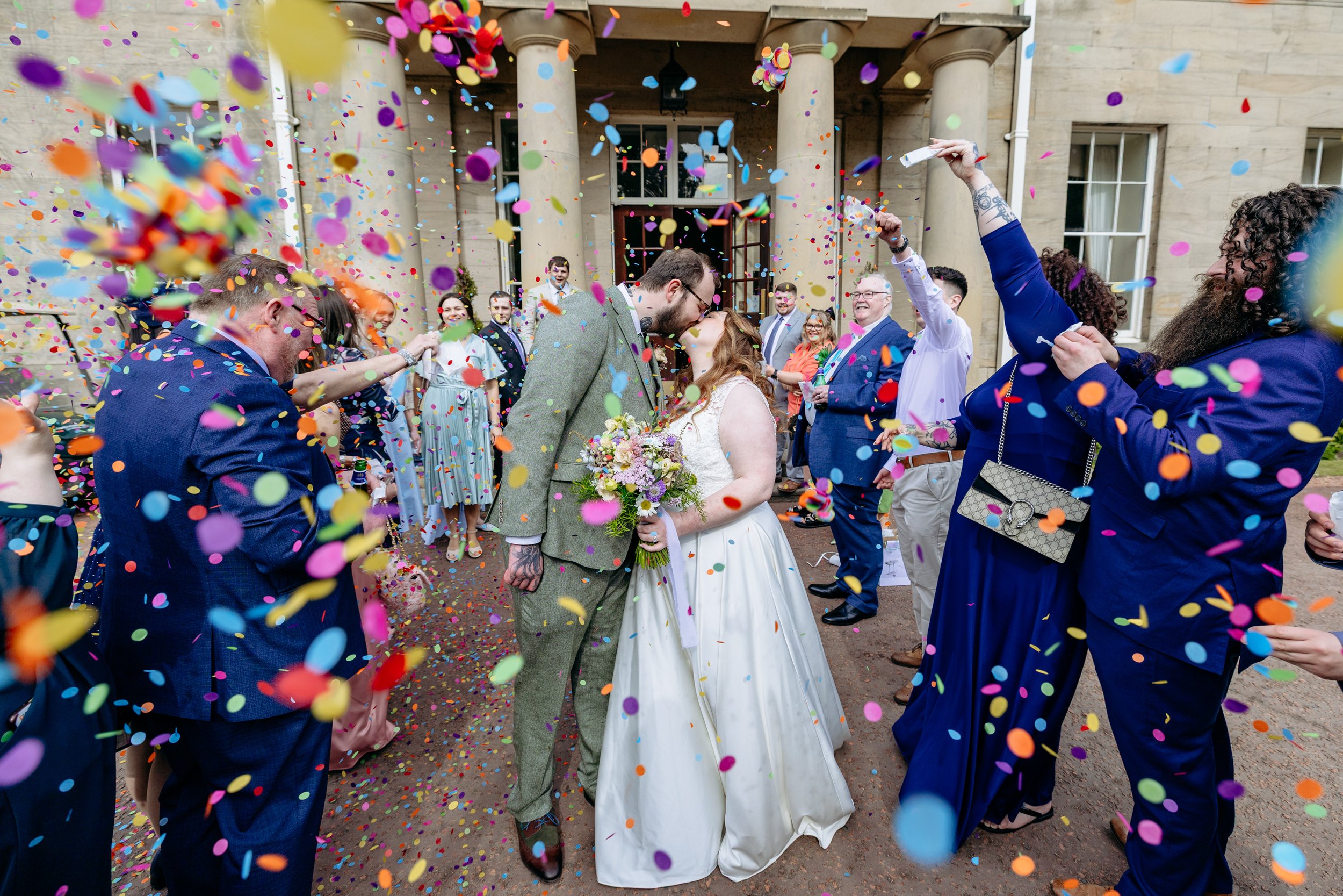 A newlywed couple sharing a kiss surrounded by wedding guests and colorful confetti outside a grand building with columns.