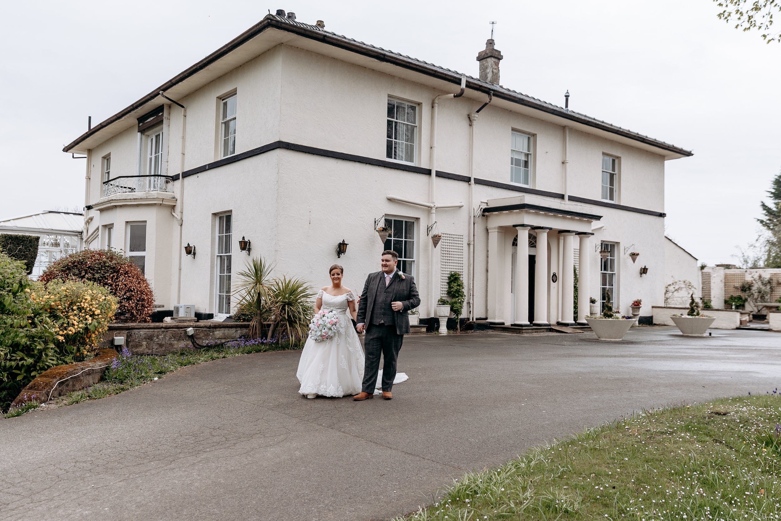 Bride in a white wedding gown holding a bouquet walking with a groom in a dark suit outside a large white house.