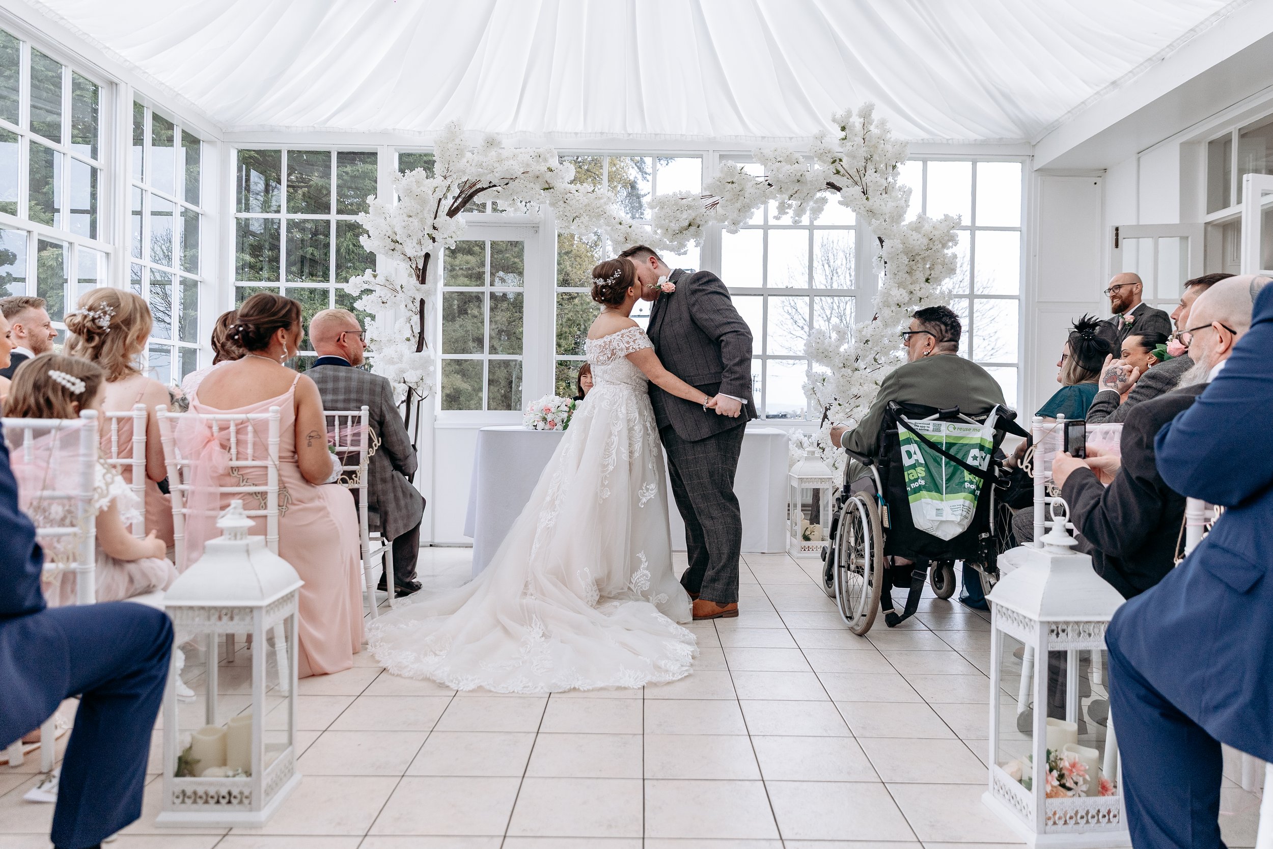Bride and groom sharing a kiss during their wedding ceremony in a sunlit conservatory decorated with white flowers, with guests seated around them.