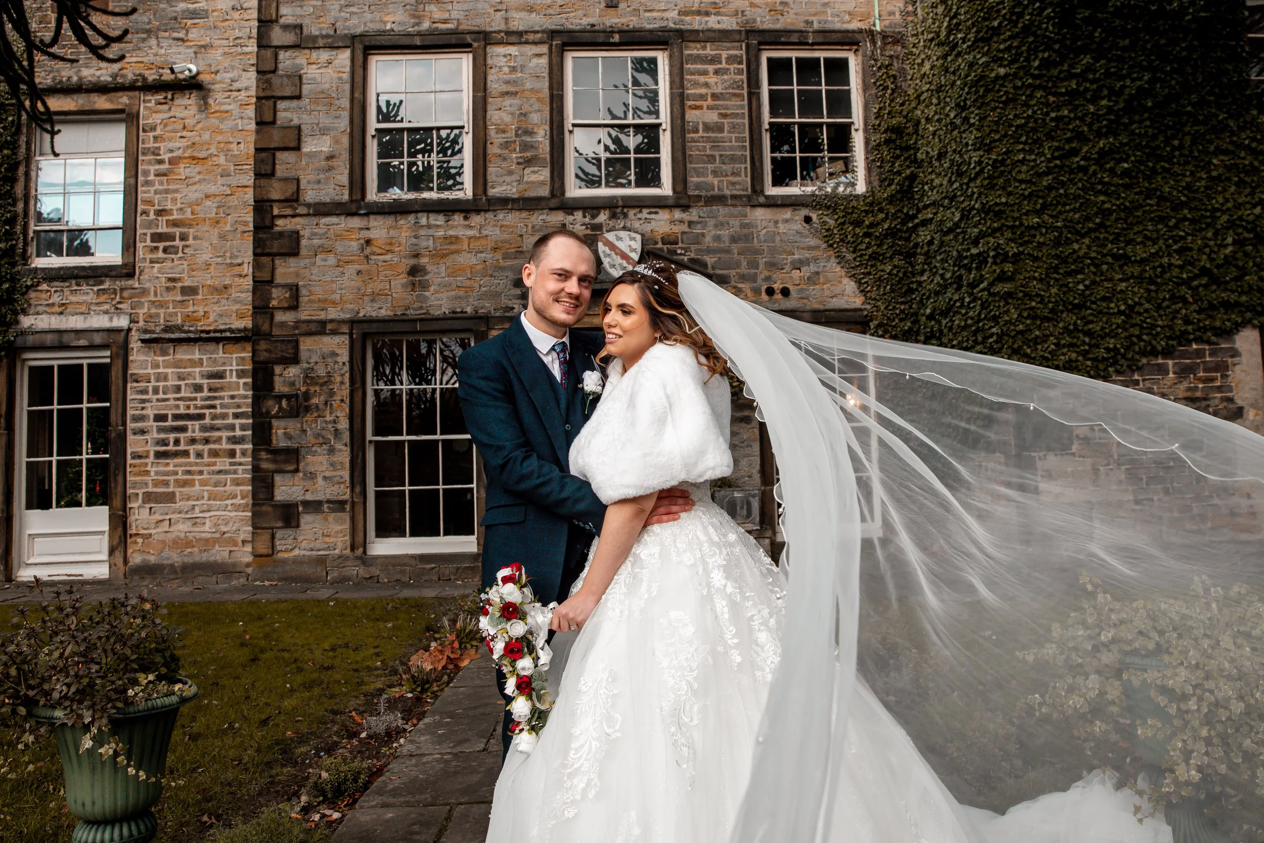 A bride and groom in wedding attire standing outdoors in front of a brick house with multiple windows. The bride is wearing a white wedding dress with a long veil and holding a bouquet, while the groom is wearing a dark suit with a tie. They are smil