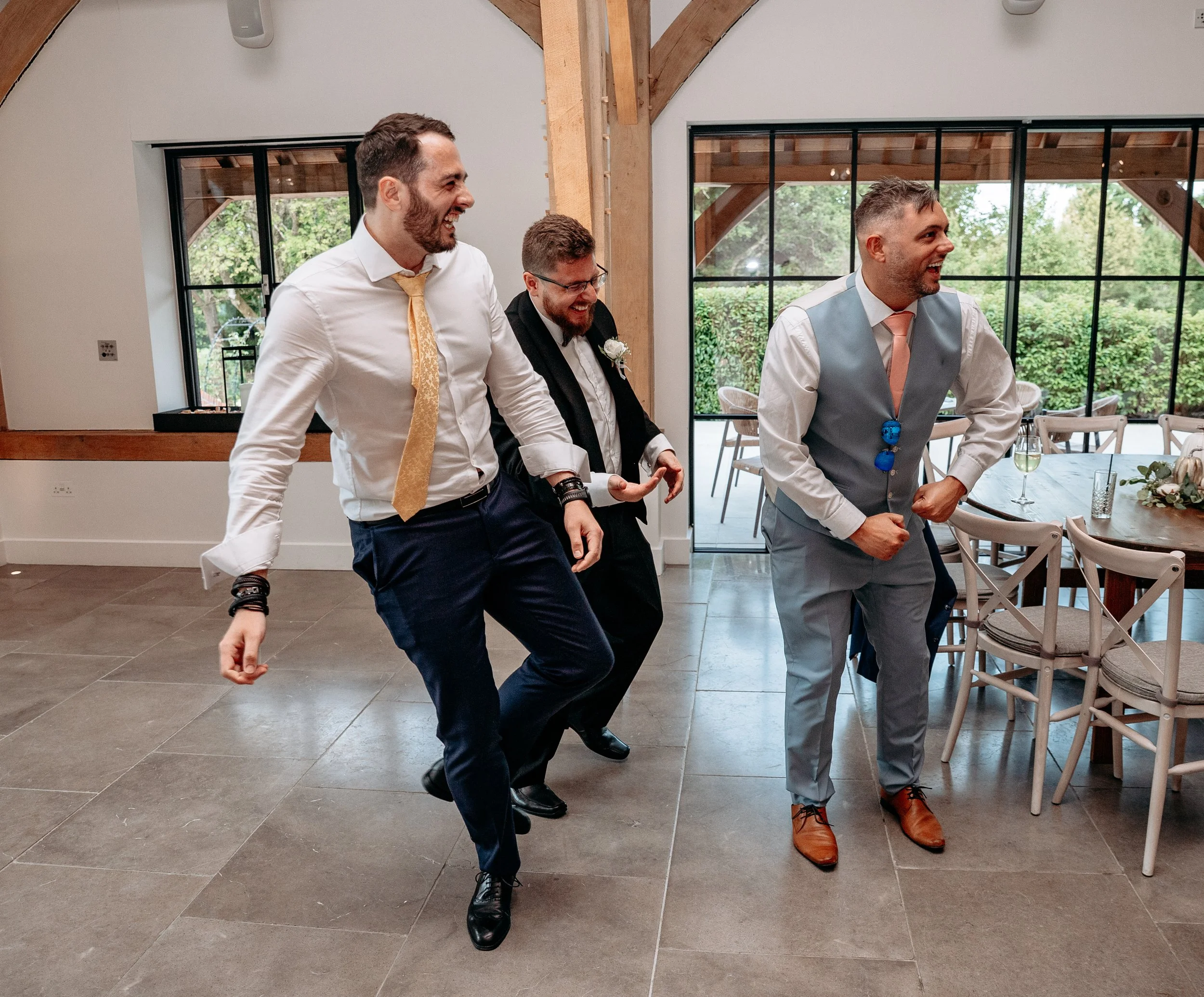 Three men in formal attire, smiling and applauding at an indoor event, possibly a wedding. The setting includes wooden beams and a large window with a view of greenery outside. A table with floral decorations is partially visible on the right.