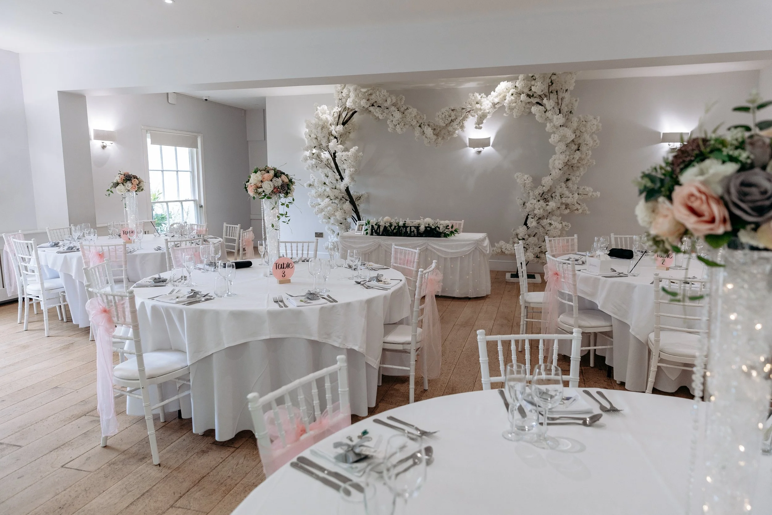 Elegant wedding reception setup with white tables, chairs, and floral centerpieces, featuring a heart-shaped floral backdrop in a decorated event space.