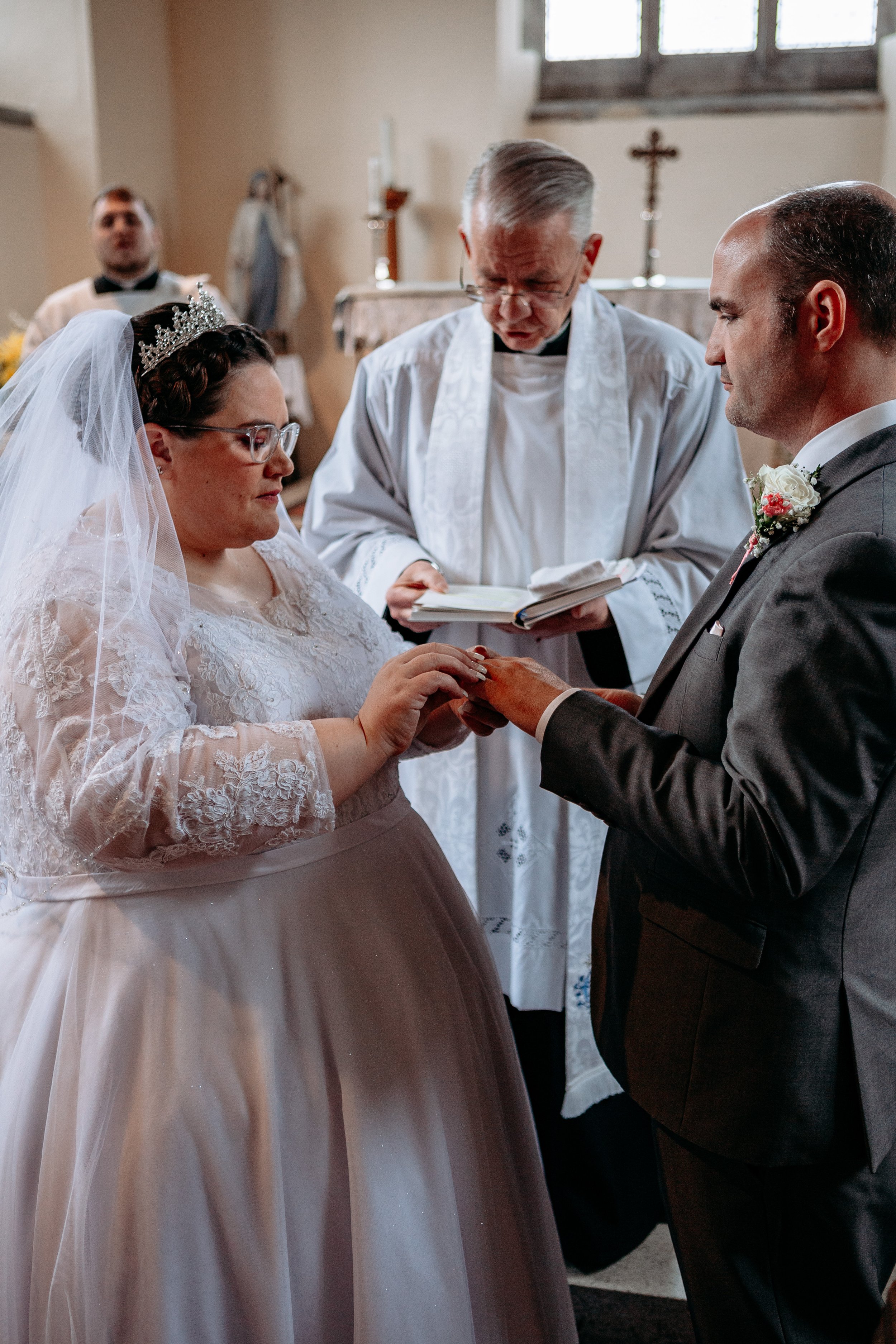 A bride and groom exchanging rings during a wedding ceremony inside a church, with an officiant reading from a book and an altar in the background.
