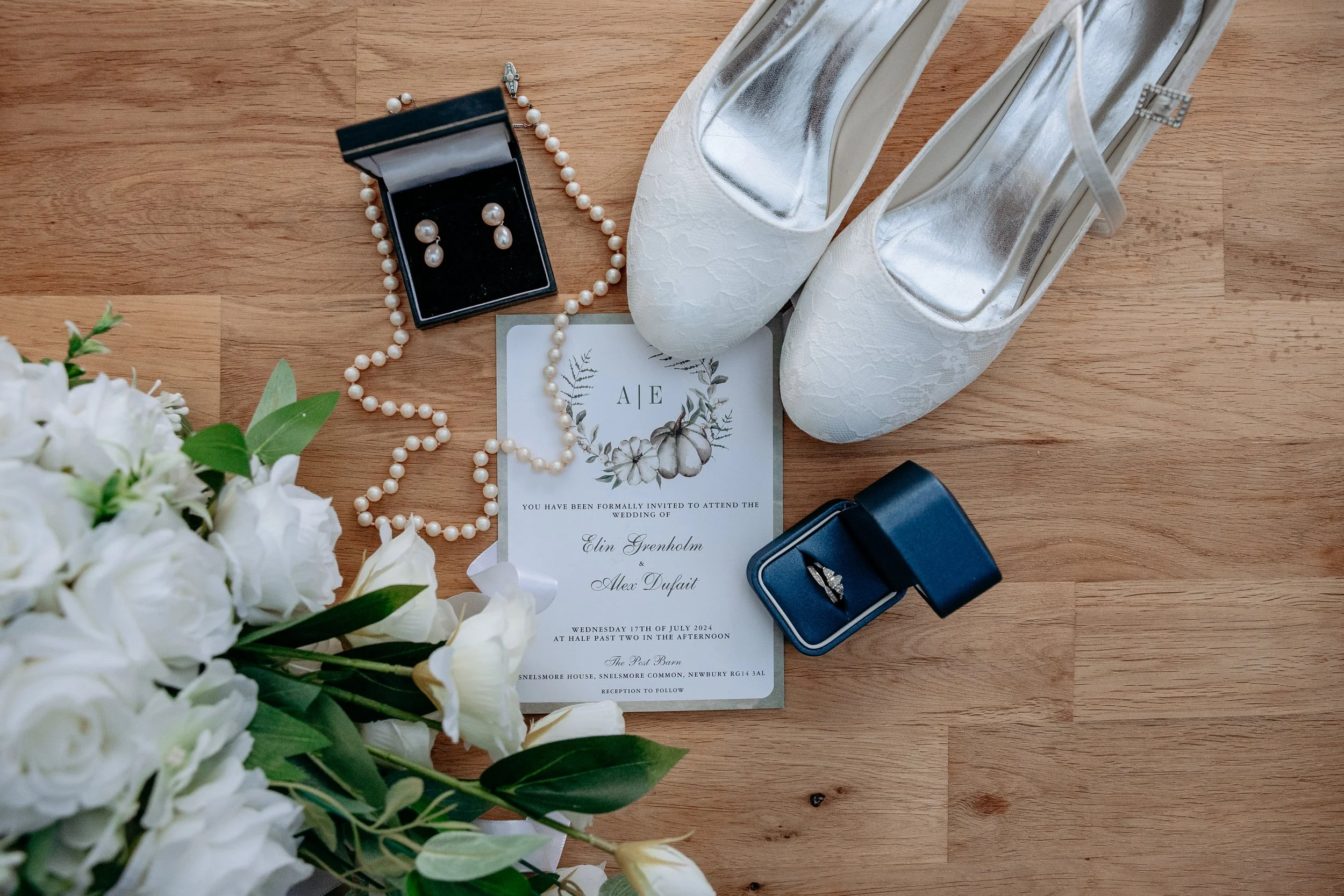 Flat lay of wedding items including white shoes, pearl necklace, earrings in a box, bouquet of white flowers, wedding invitation, and a ring in a blue box on a wooden surface.