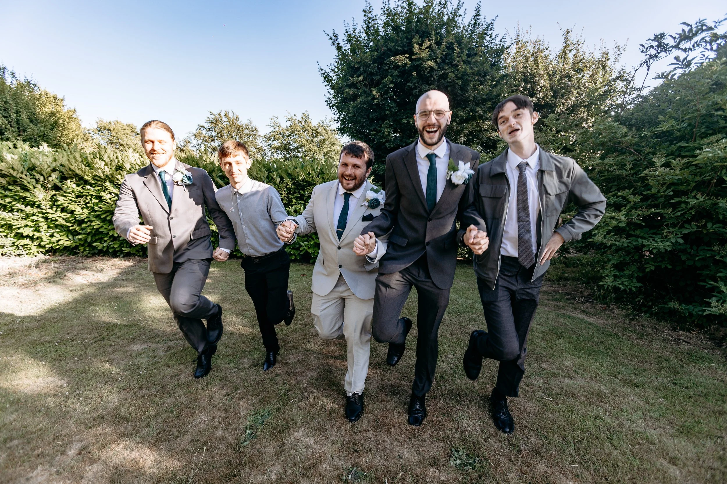 Group of five men running outdoors in a wedding or formal event, dressed in suits and ties, some with boutonnières, holding hands and smiling.
