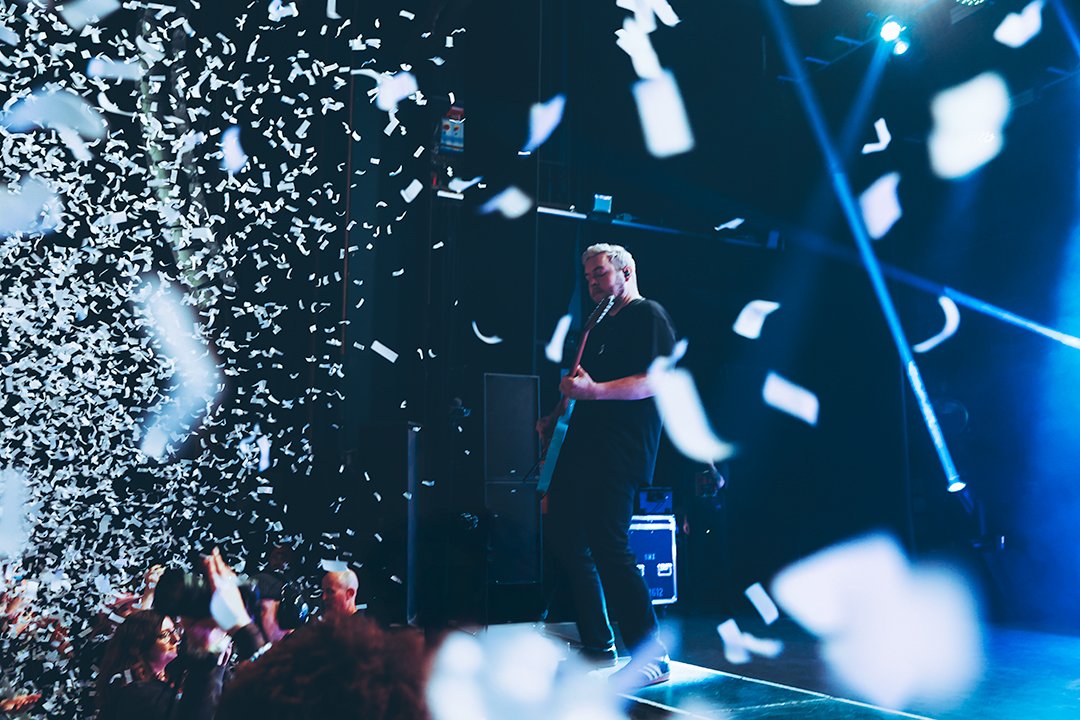 Musician playing guitar on stage with confetti falling in a concert setting, audience visible in the foreground.
