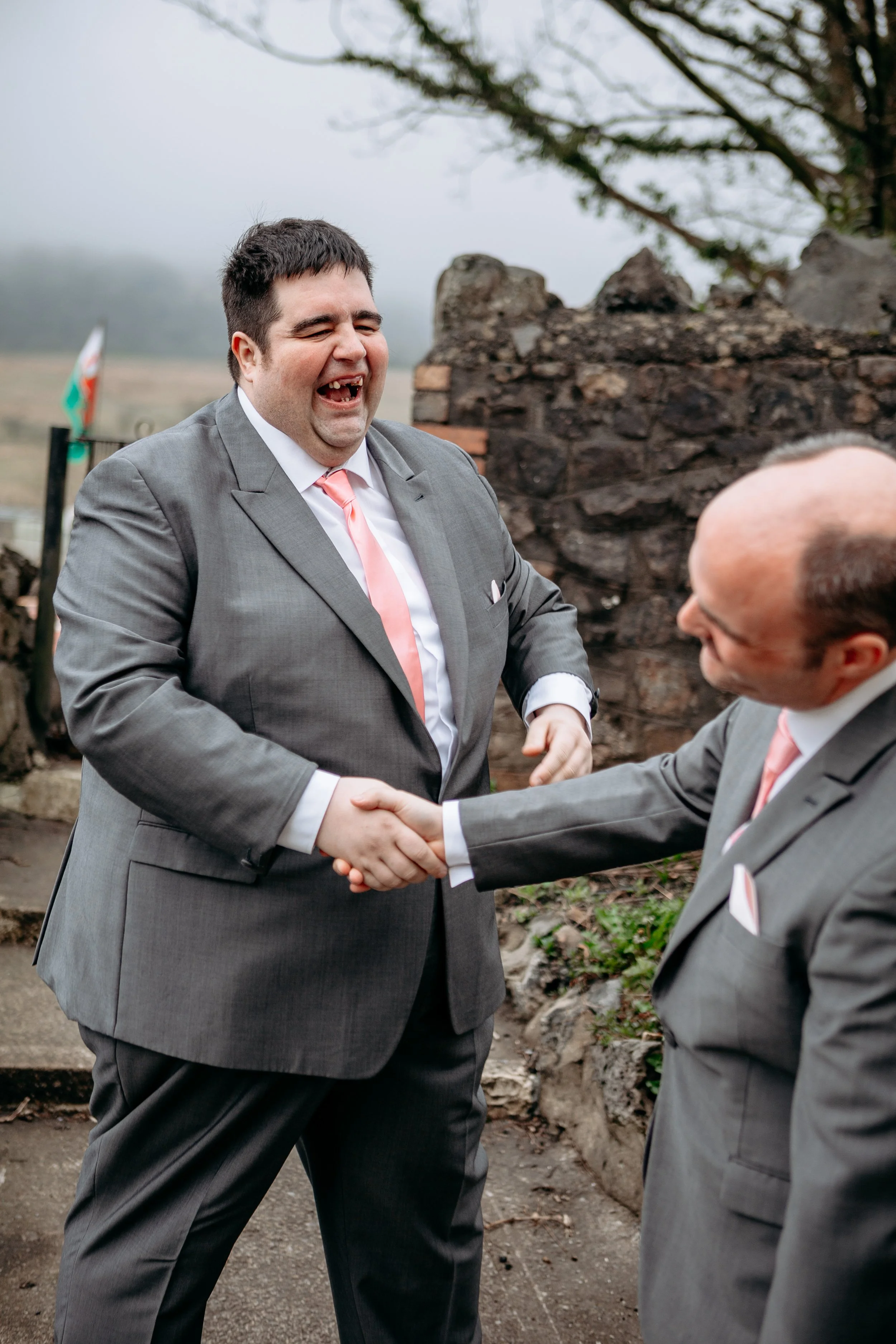 Two men in gray suits and pink ties shaking hands outdoors, smiling and laughing, near a stone wall and leafless trees.