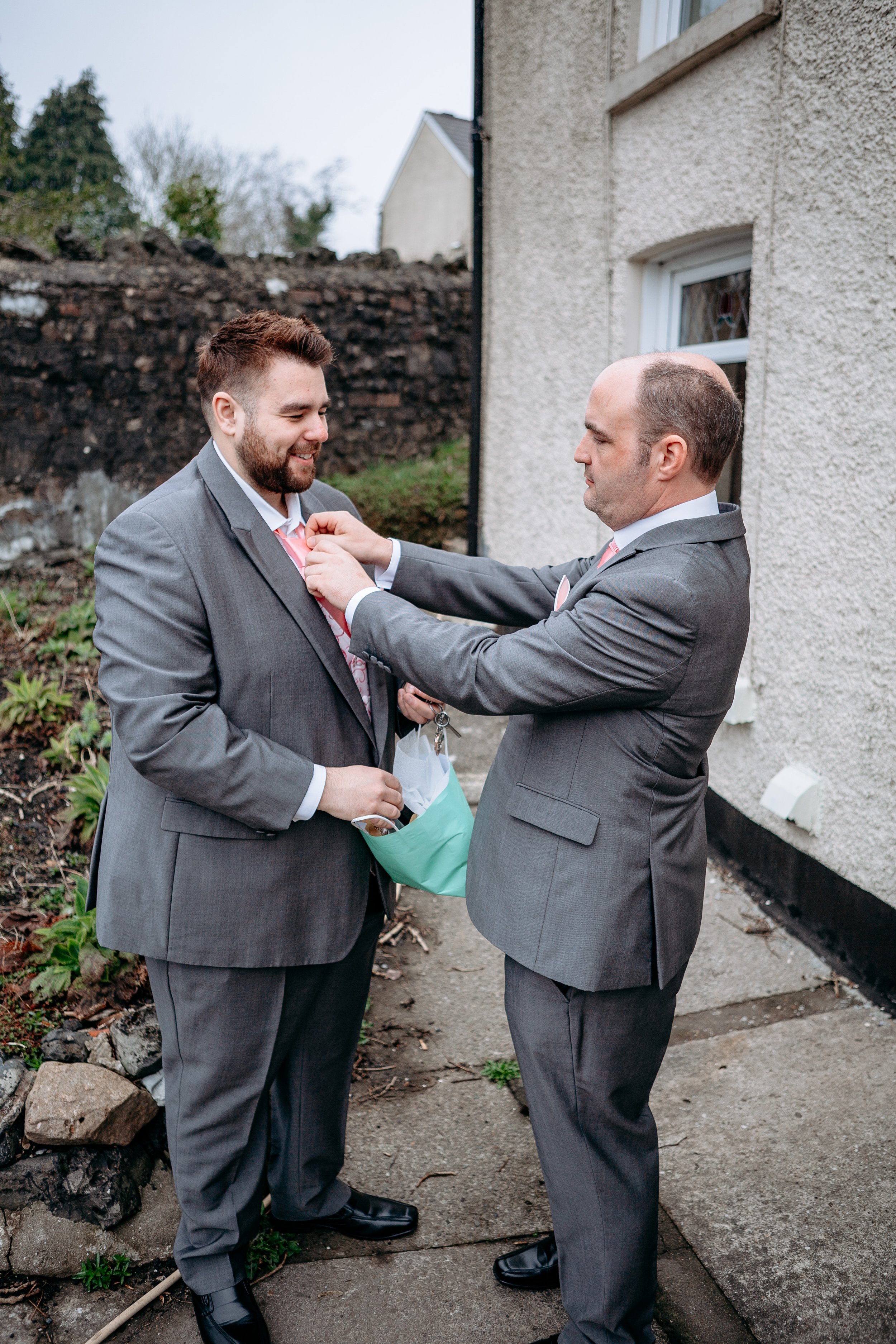 Two men in gray suits, one pinning a boutonniere onto the other's lapel outside a house.