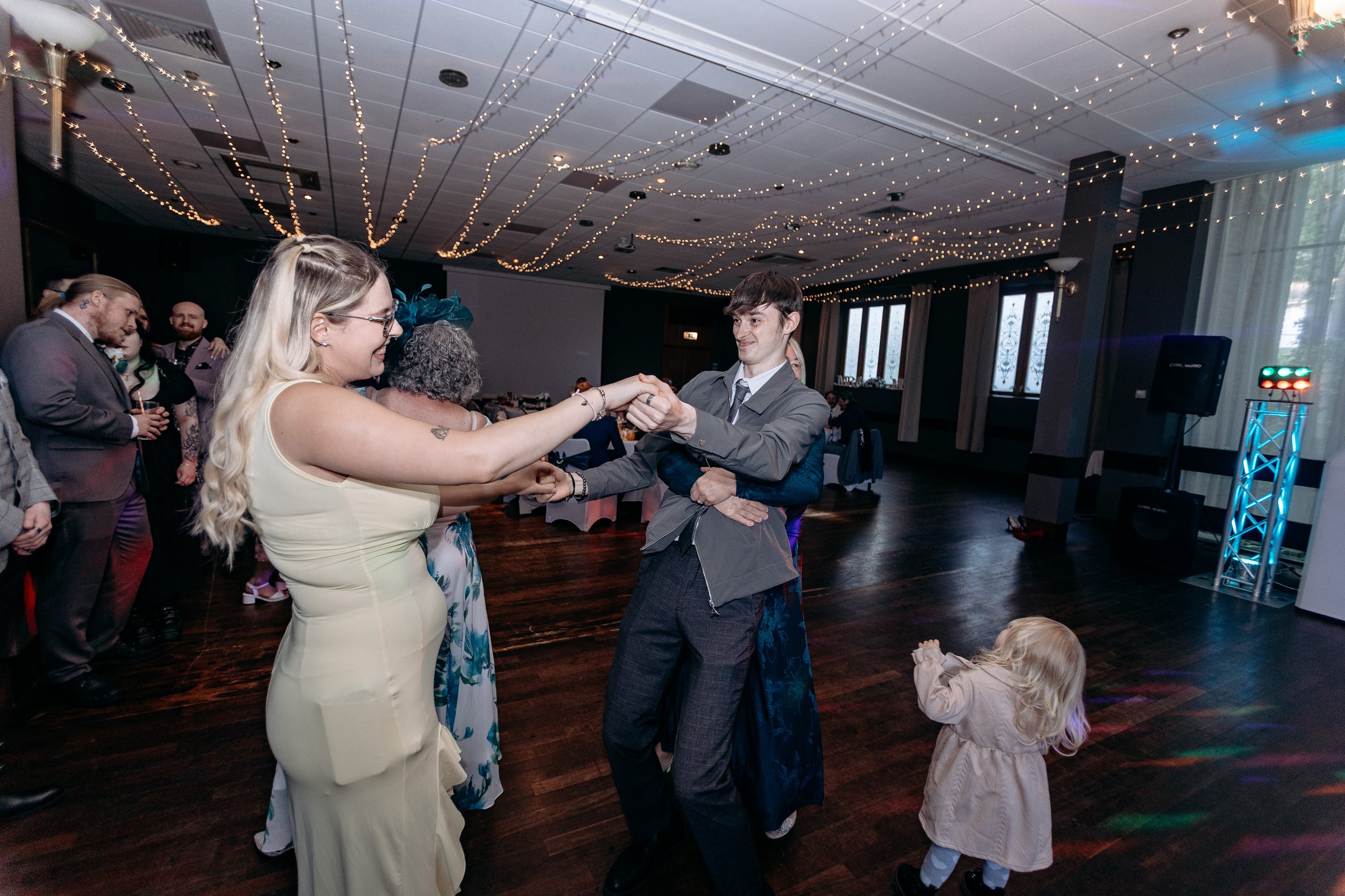 People dancing at a celebration or wedding reception with string lights hanging from the ceiling and a wooden floor.