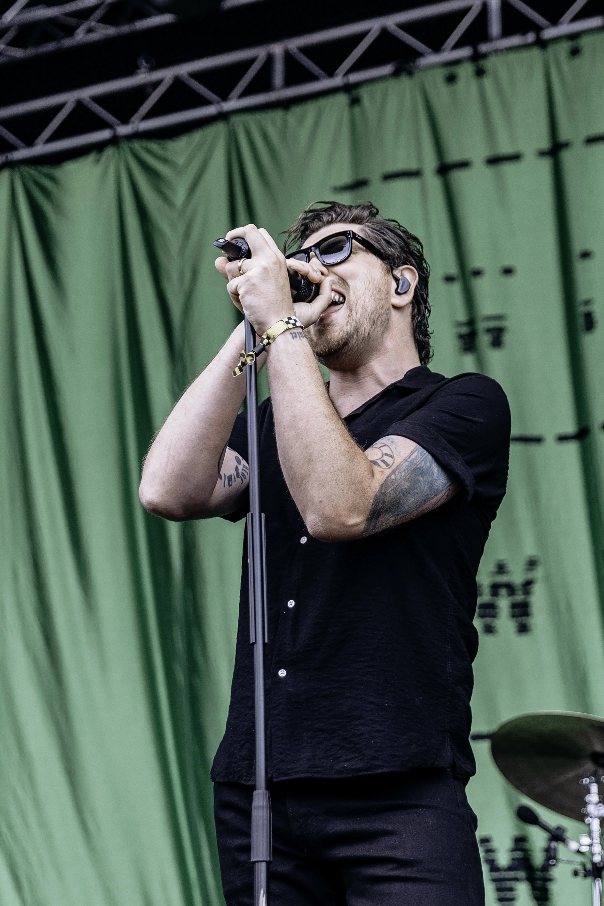 A man with curly hair, wearing sunglasses and a black shirt, sings into a microphone on stage, with a green backdrop and drums in the background.