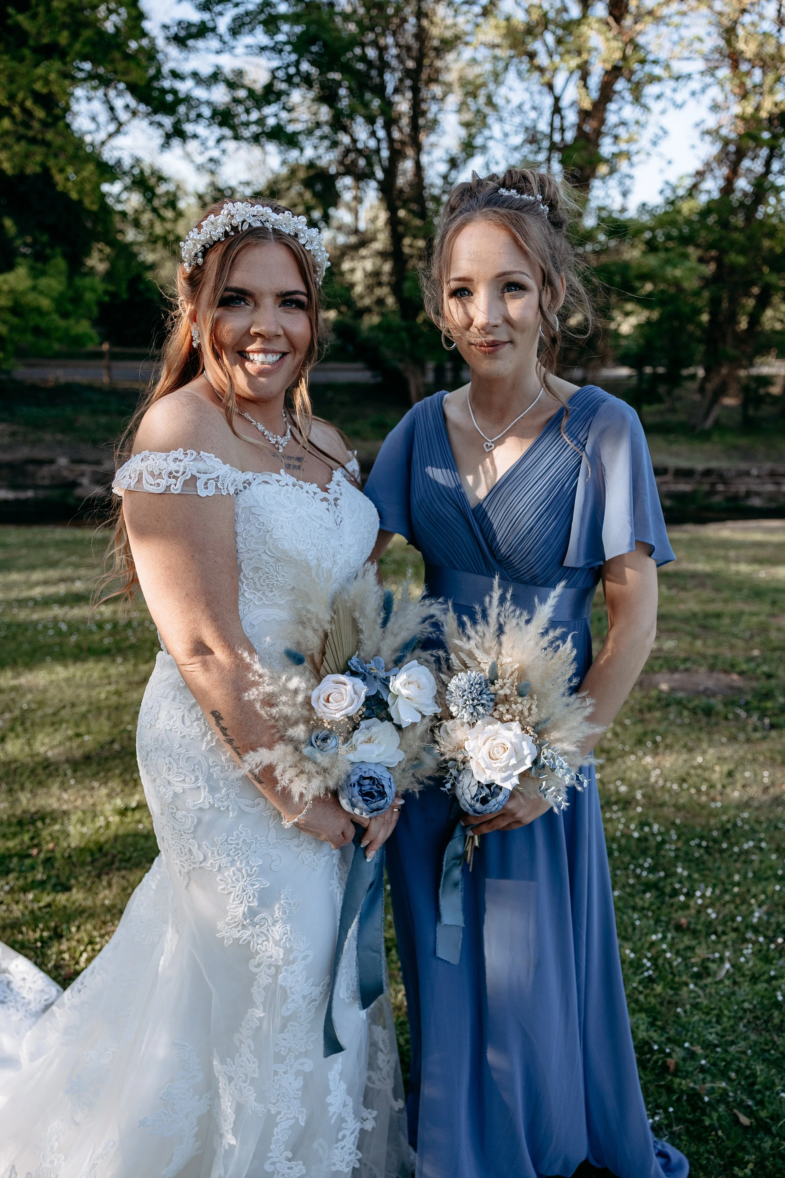 Two women standing outdoors, one in a white wedding dress with lace and off-the-shoulder sleeves, holding a bouquet of white and blue flowers with pampas grass, and the other in a blue dress holding a similar bouquet. They are smiling and posing for 