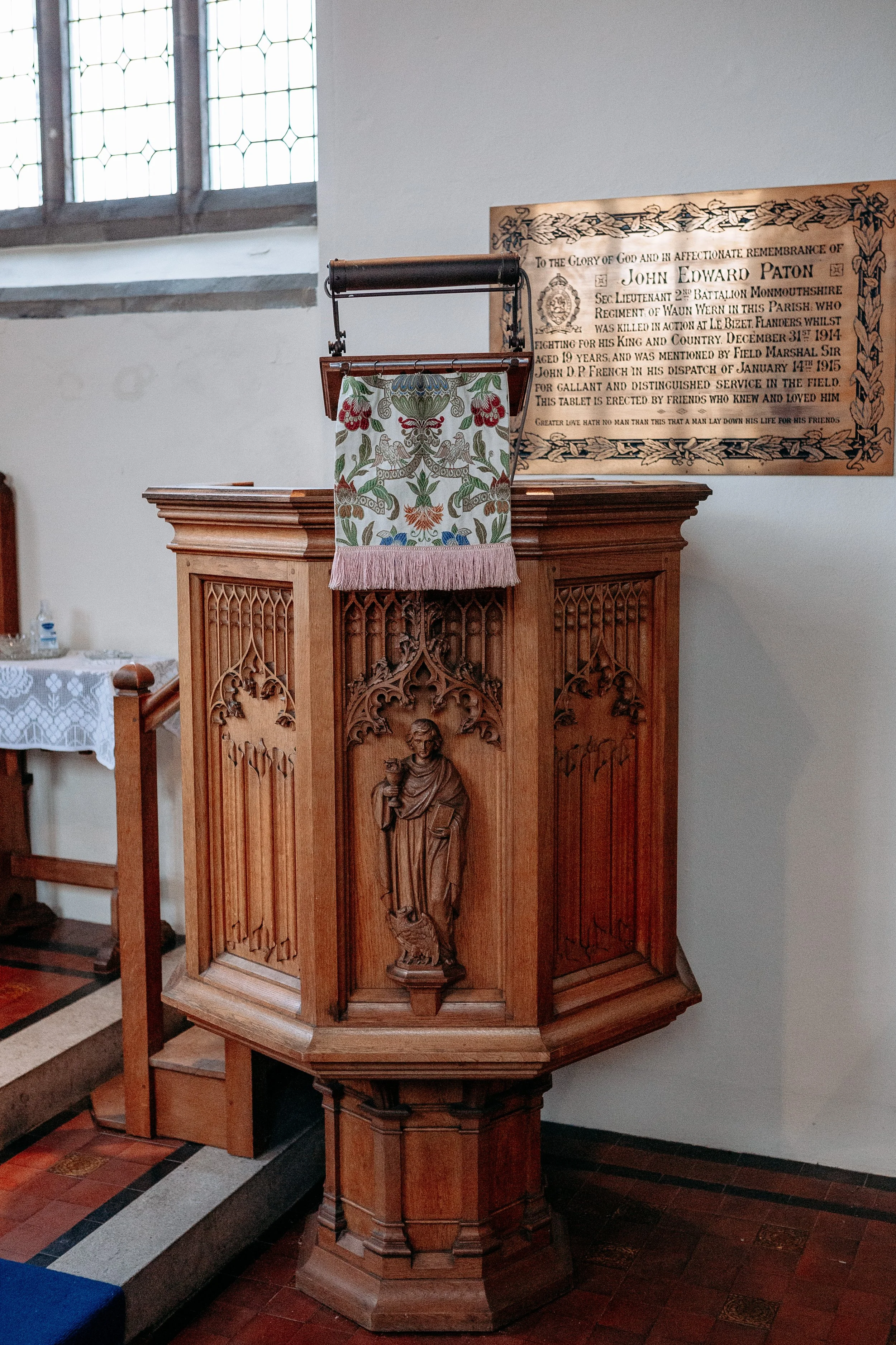 Wooden pulpit in a church with a carved figure of a saint, an embroidered cloth hanging from a brass bar, a plaque with engraved text, and stained glass windows in the background.