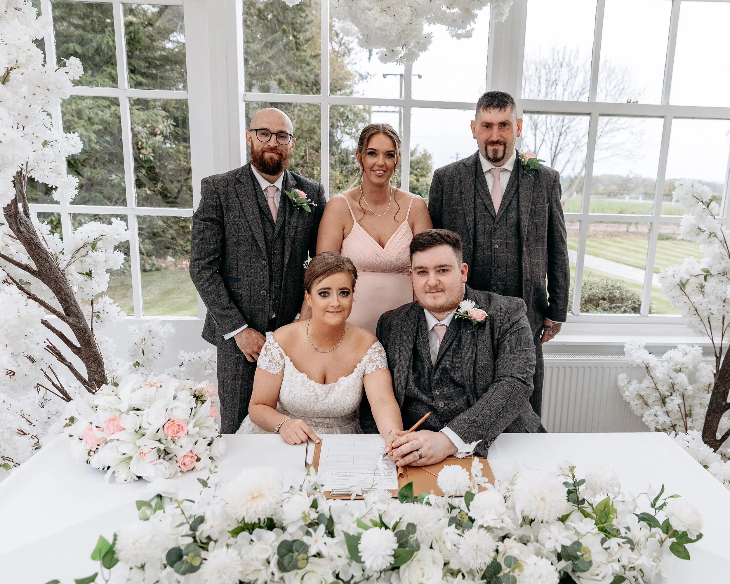 A wedding party of six people, four men and two women, gathered around a table with wedding documents and flowers, inside a bright room with large windows revealing trees and a cloudy sky outside.