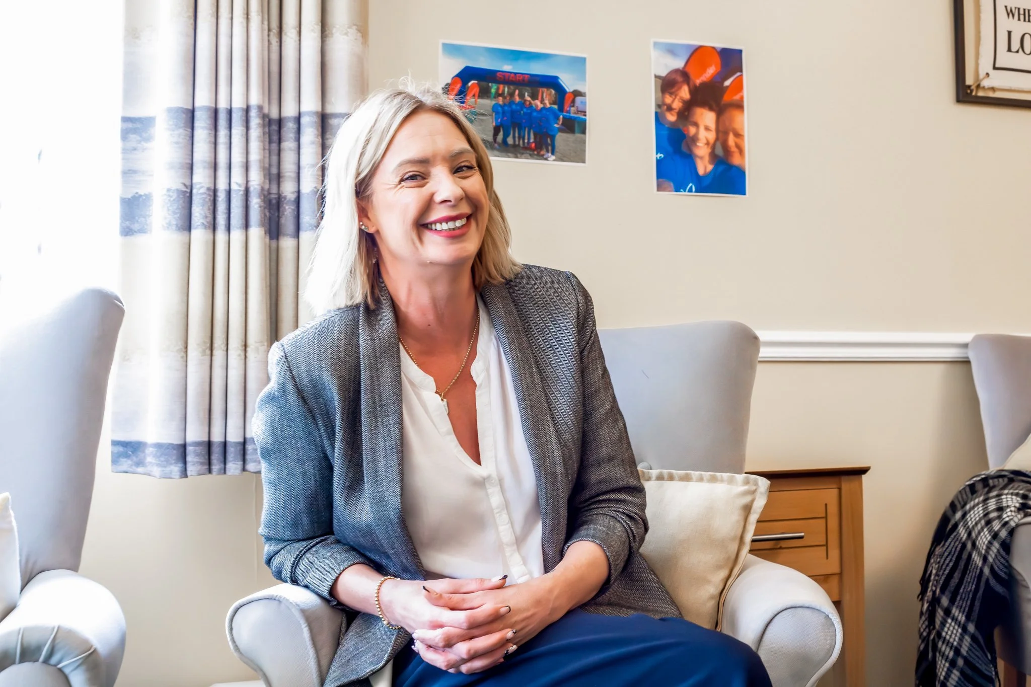 A smiling woman with shoulder-length blonde hair sitting on a white armchair in a living room, wearing a gray blazer and white blouse, with photos on the wall behind her.