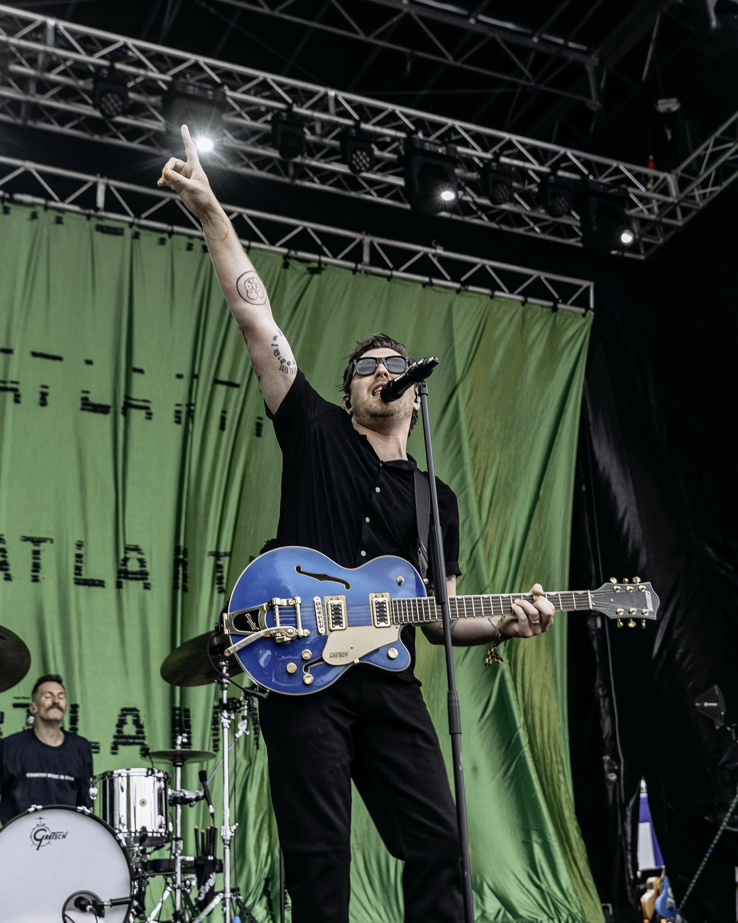 A musician with sunglasses sings into a microphone while playing a blue electric guitar on stage during a live performance.