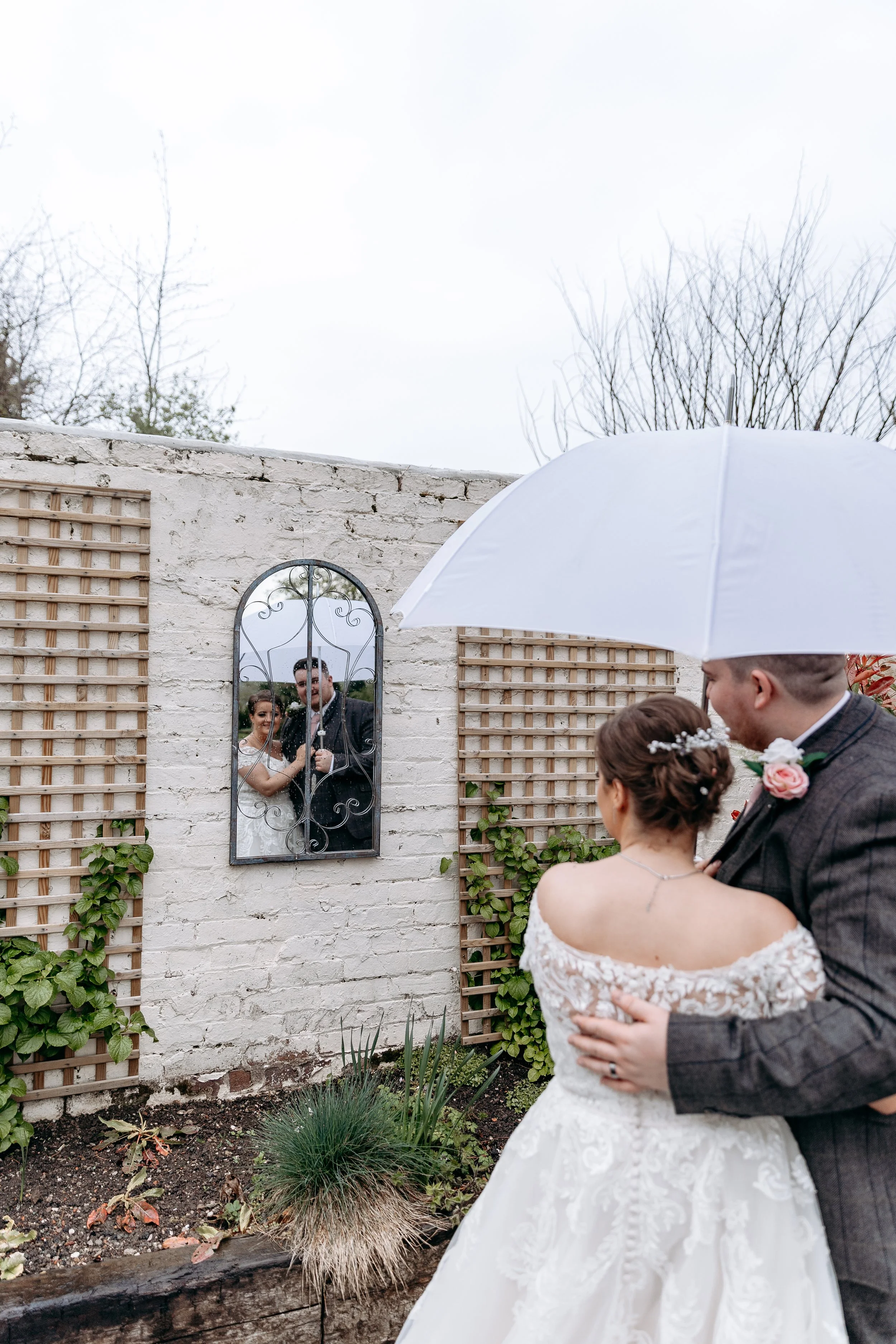 A newlywed couple sharing a romantic moment outdoors under an umbrella, looking at their reflection in a mirror mounted on a white brick wall, on a cloudy day with leafless trees in the background.