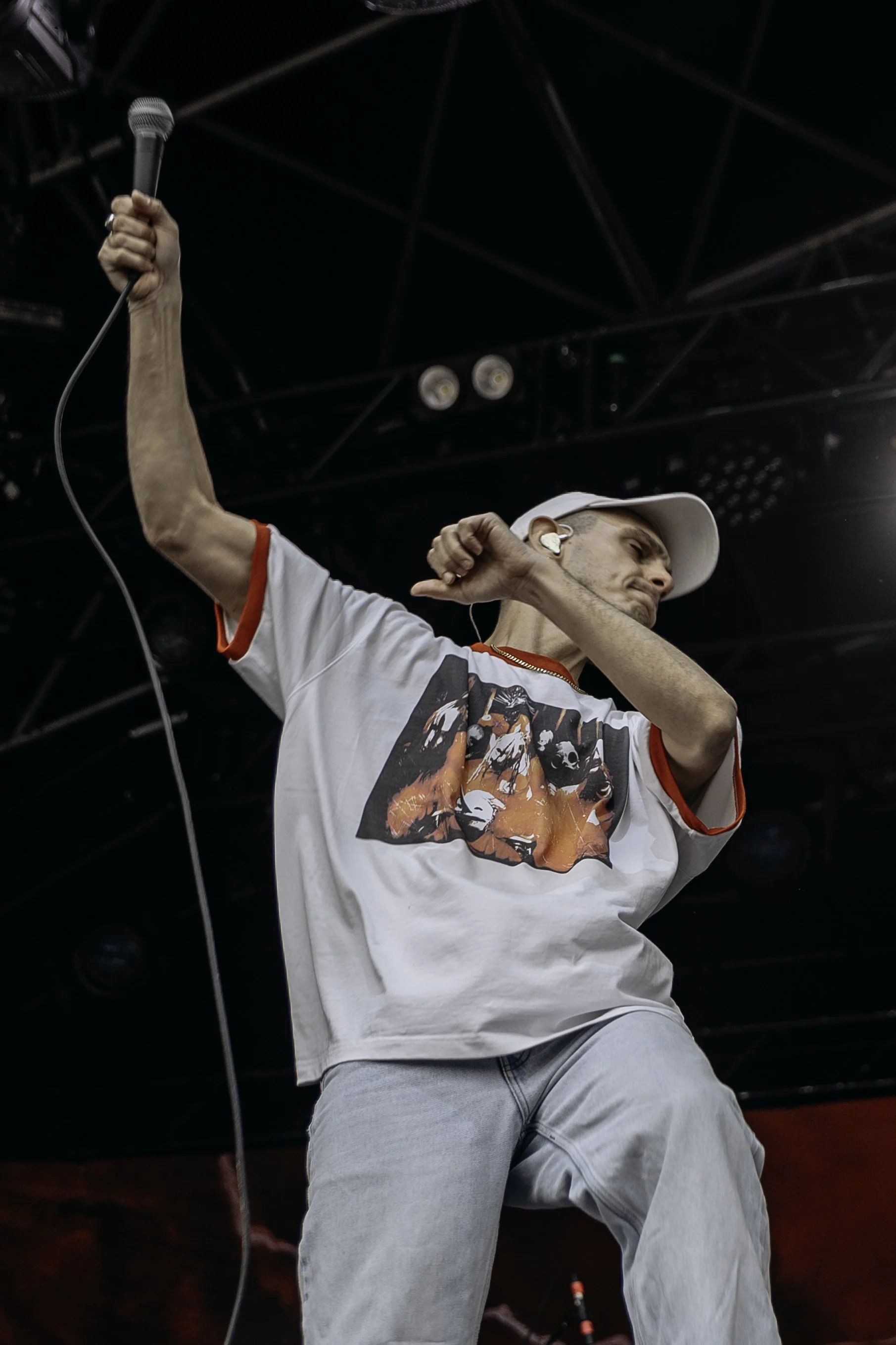 A male performer on stage holding a microphone in one hand while dancing, wearing a white t-shirt with a graphic design, light blue jeans, a white cap, and earphones in a dark concert venue.