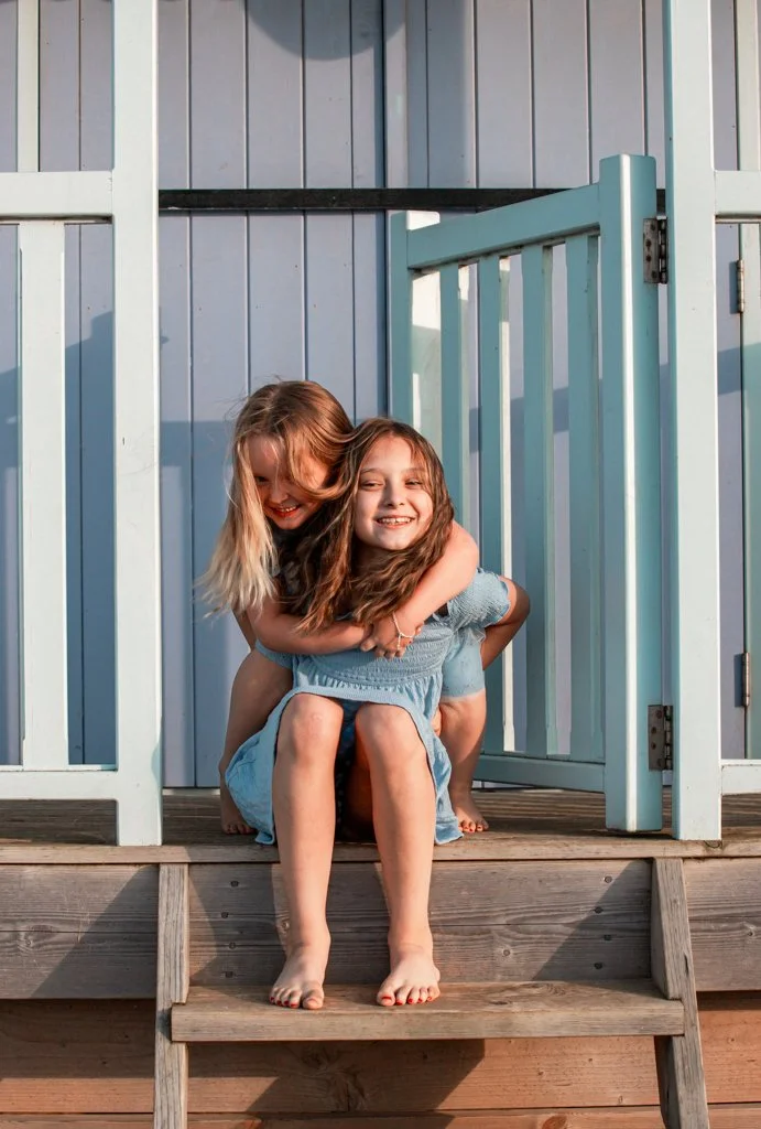 Two smiling girls sitting on wooden steps in front of a light blue building, playing and embracing each other.