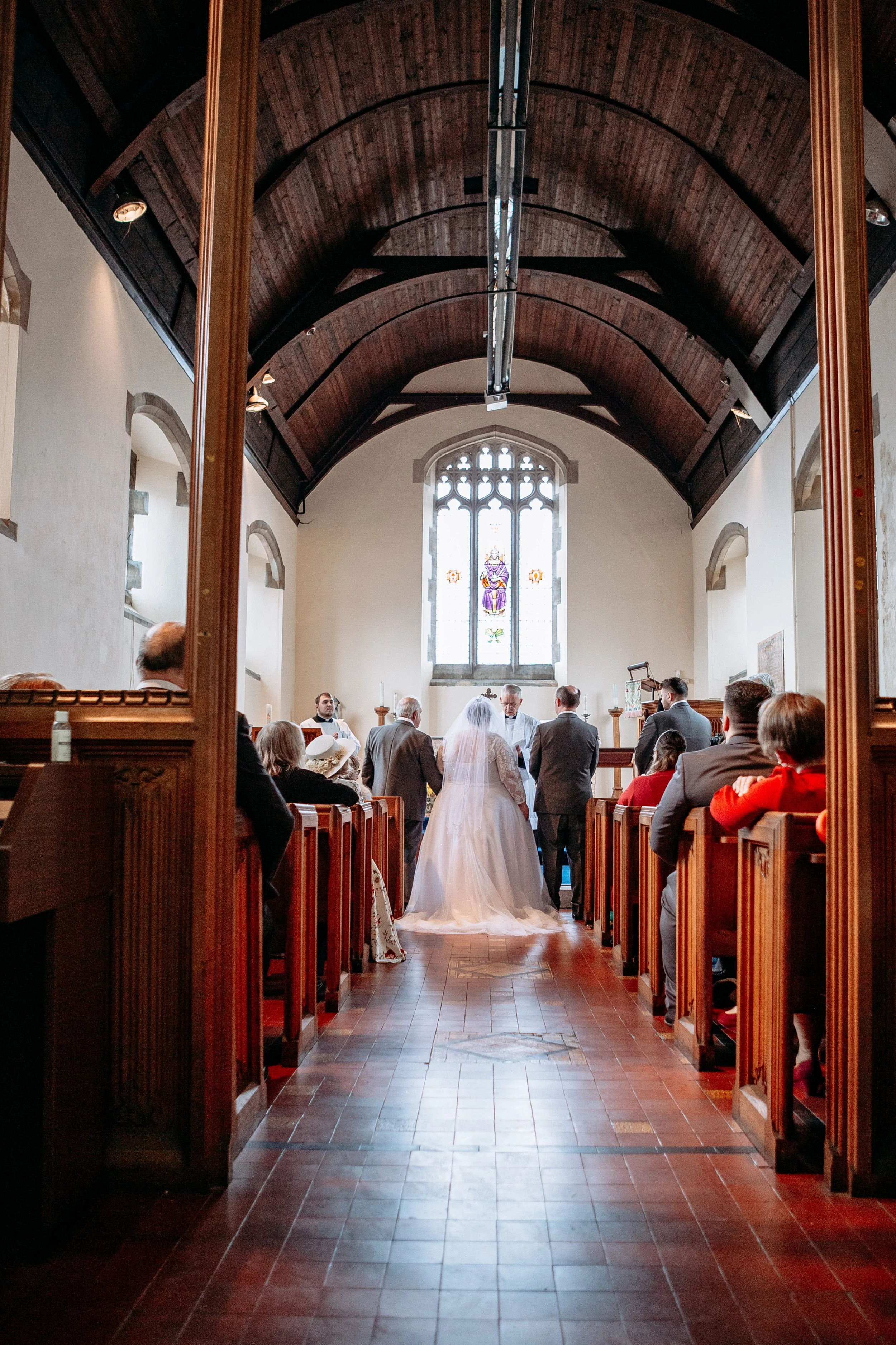 A wedding ceremony inside a small church with a priest, bride in a white gown, groom, and guests seated on wooden pews, with a stained glass window behind the altar.
