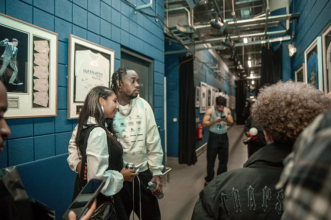 People standing in a hallway with framed memorabilia on the walls, including autographed shirts and photos. The setting appears to be backstage or a behind-the-scenes area.