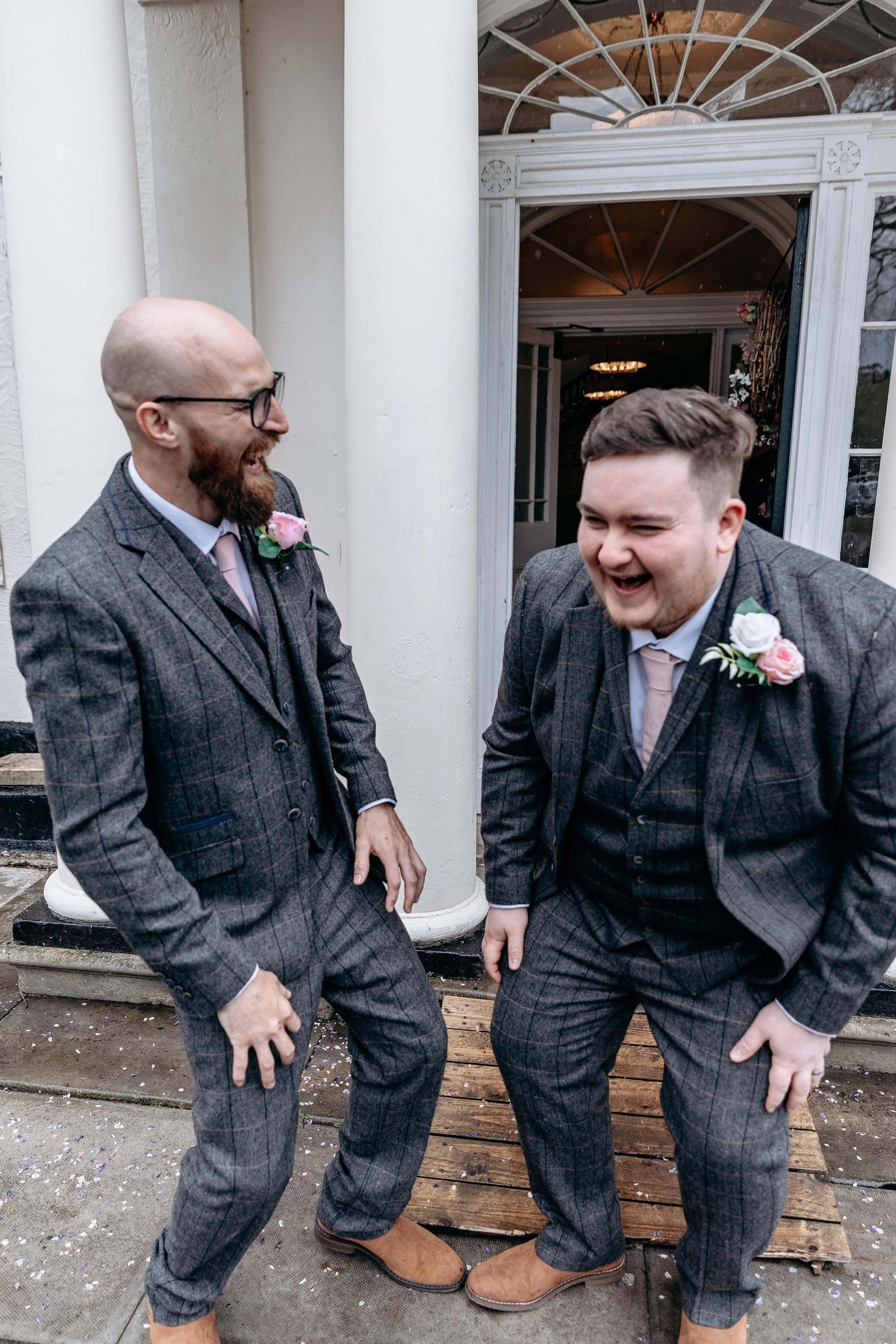 Two men in matching gray plaid suits and brown shoes standing outside a building with white columns, laughing and talking, with confetti on the ground.