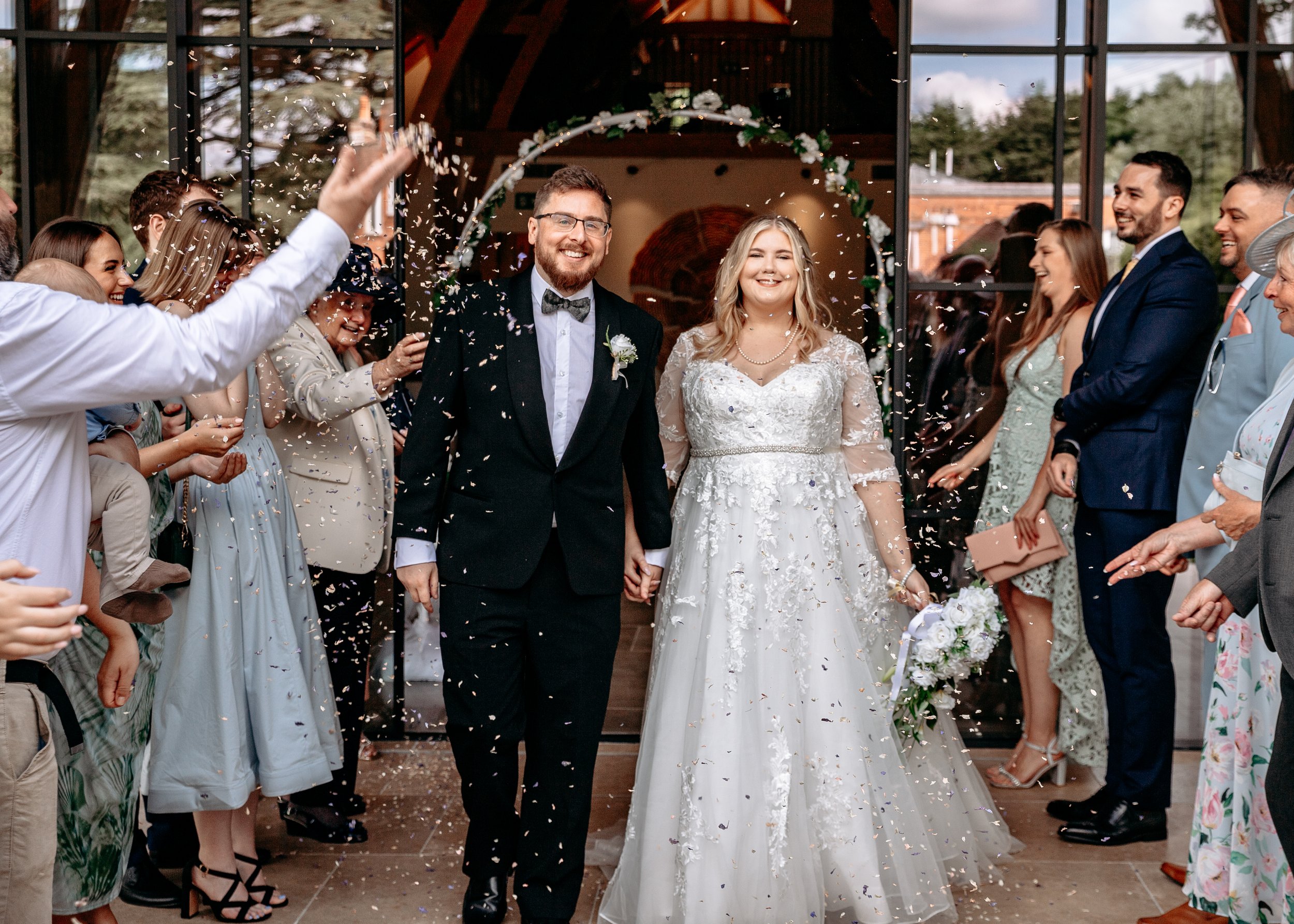 Bride and groom walking down a confetti-lined aisle with guests cheering, at a wedding ceremony.