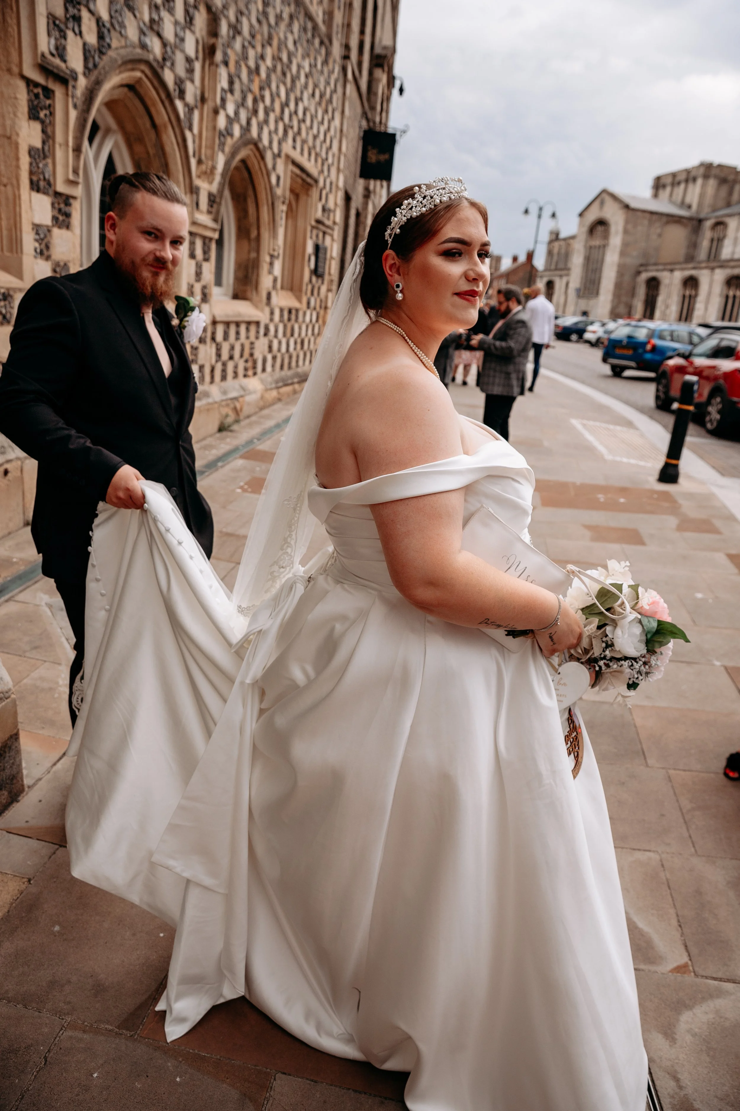 A bride in a white off-the-shoulder wedding dress holding a bouquet, and a groom in a black suit holding her dress train, standing outside a historic building on a cloudy day.