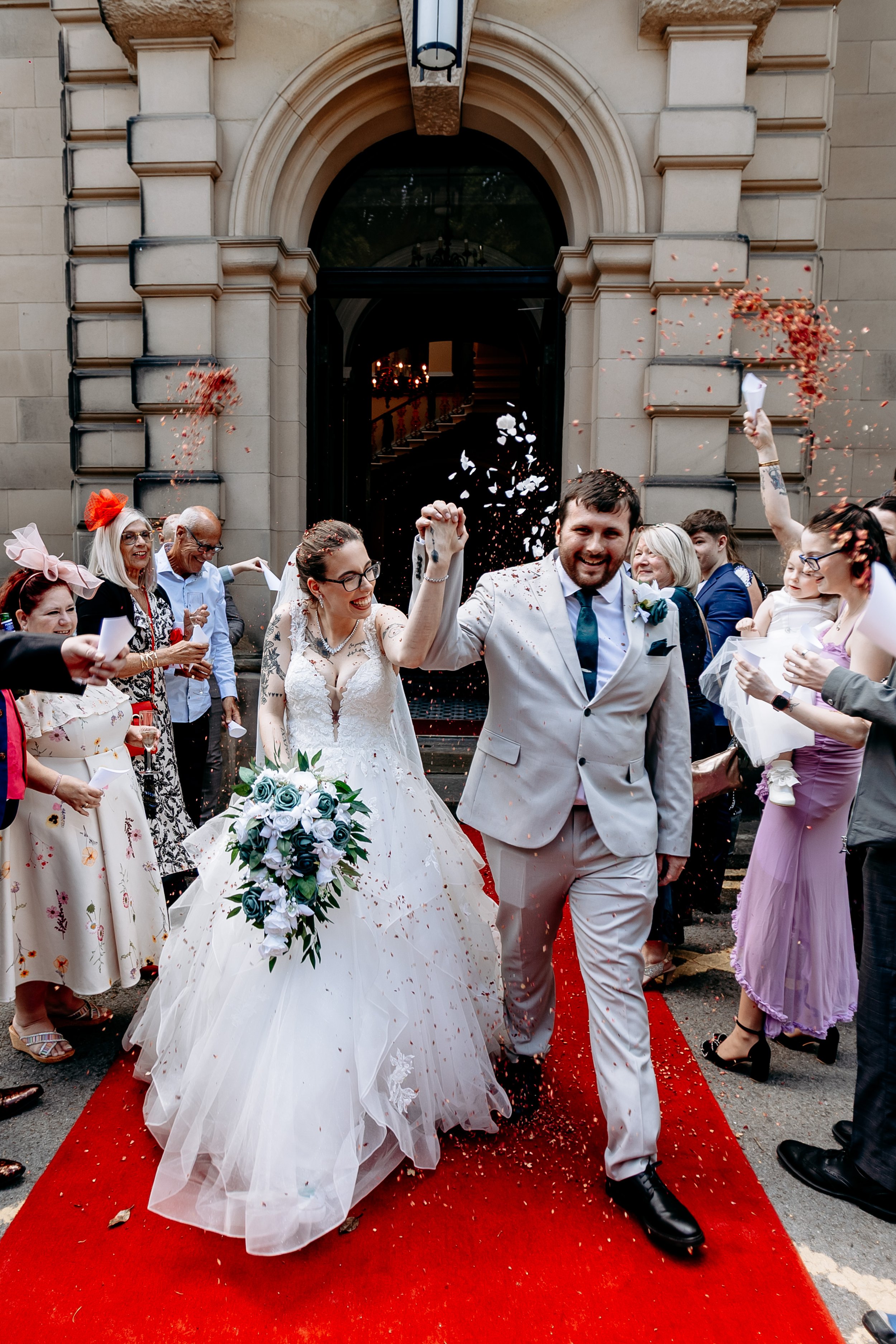 A newlywed couple walking down a red carpet outside a building, celebrating with guests throwing confetti, with the bride holding a bouquet of white and blue roses and the groom in a light-colored suit.