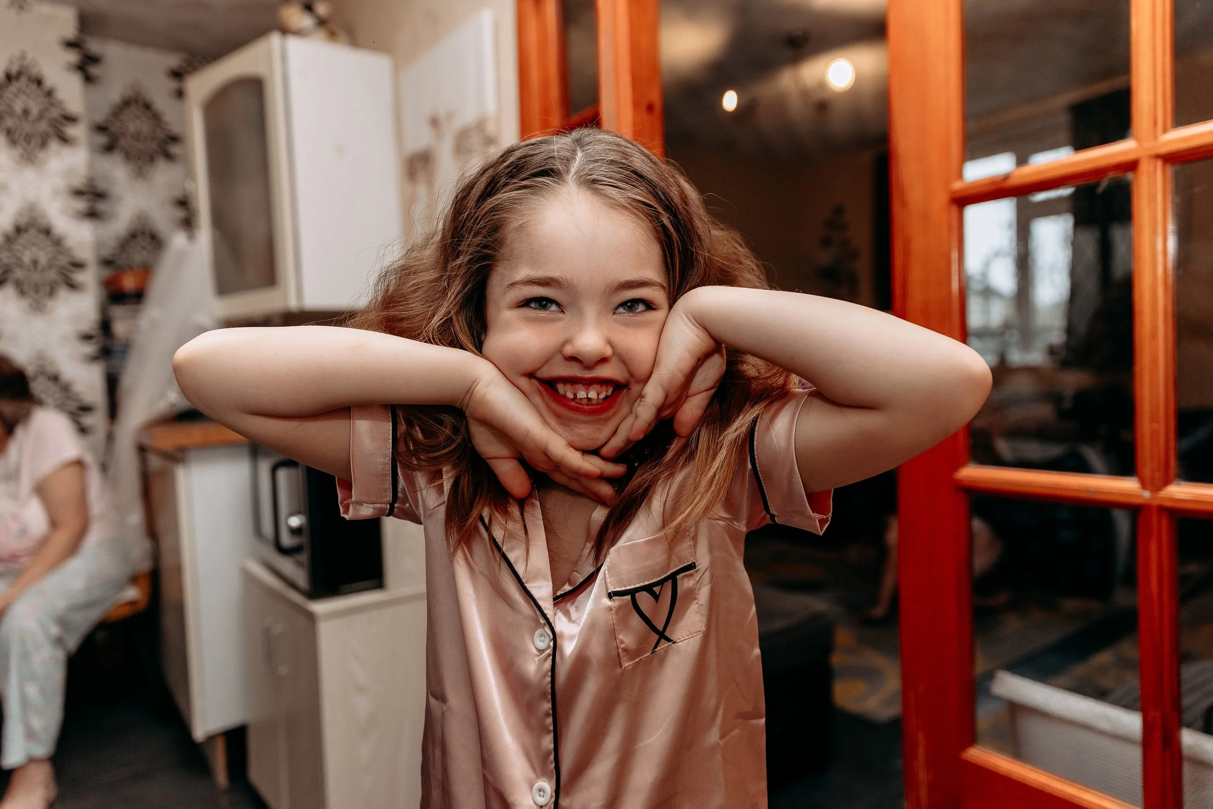 A young girl with light brown hair and blue eyes smiling and making a playful face, resting her chin on her hands, in a cozy home setting.
