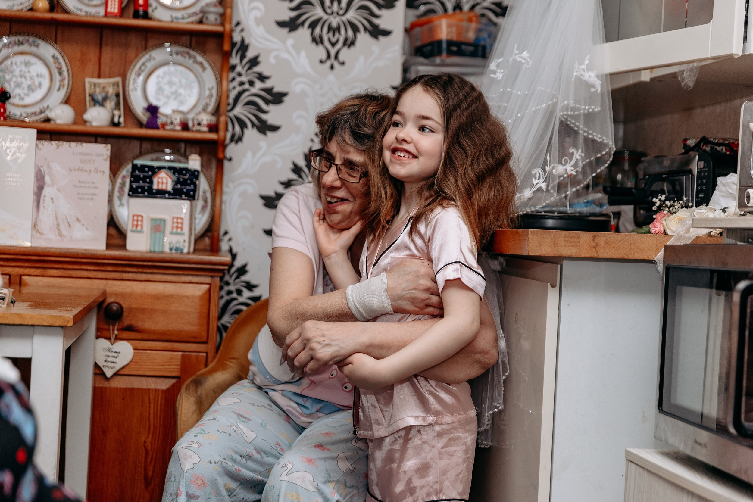 A woman and a young girl, both in pajamas, embrace and smile in a kitchen decorated for Christmas with festive plates and cards on the wall behind them.