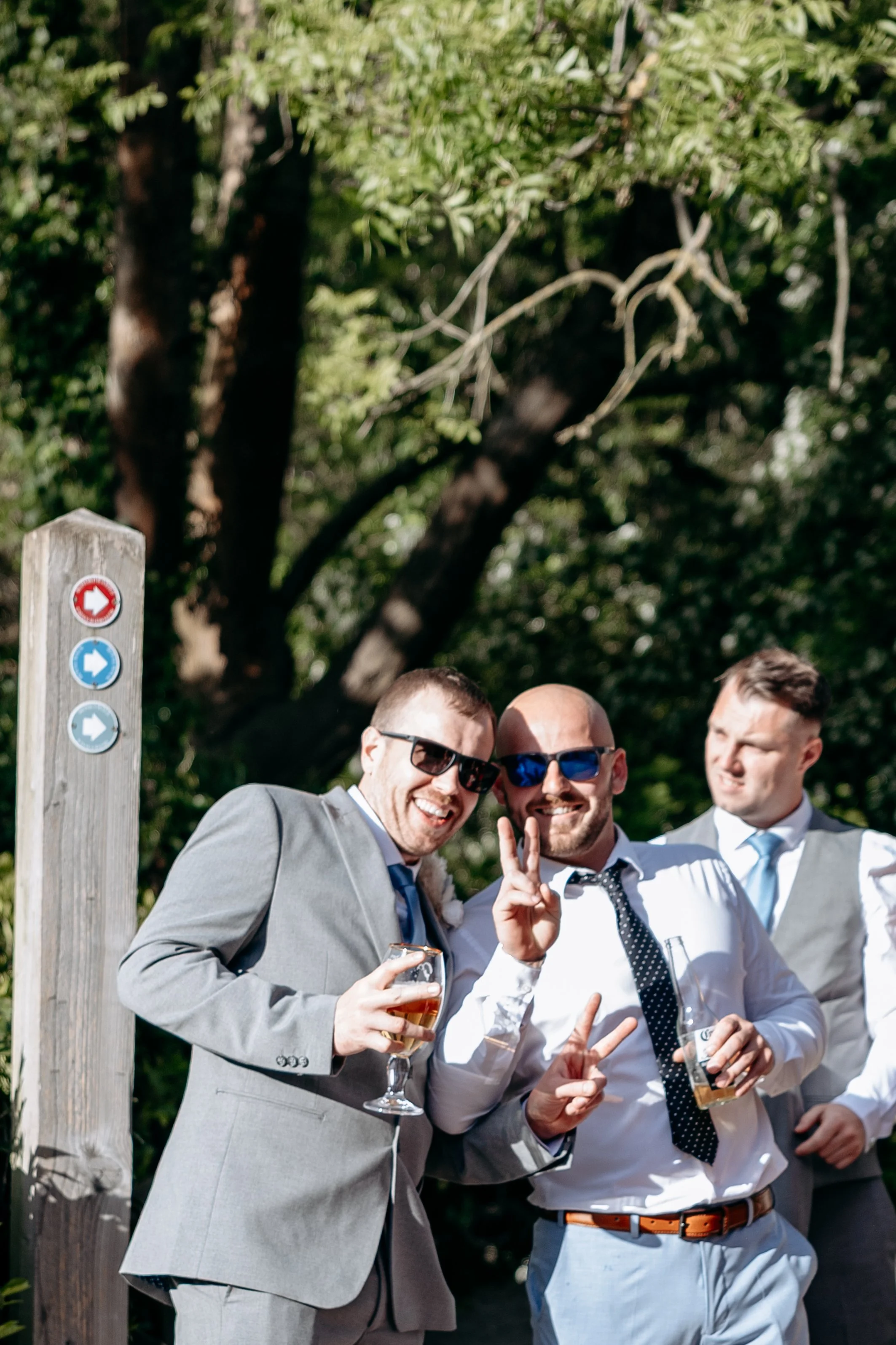 Three men wearing suits and sunglasses at an outdoor event, smiling and holding drinks, with a wooded background and a wooden post with colorful directional signs.