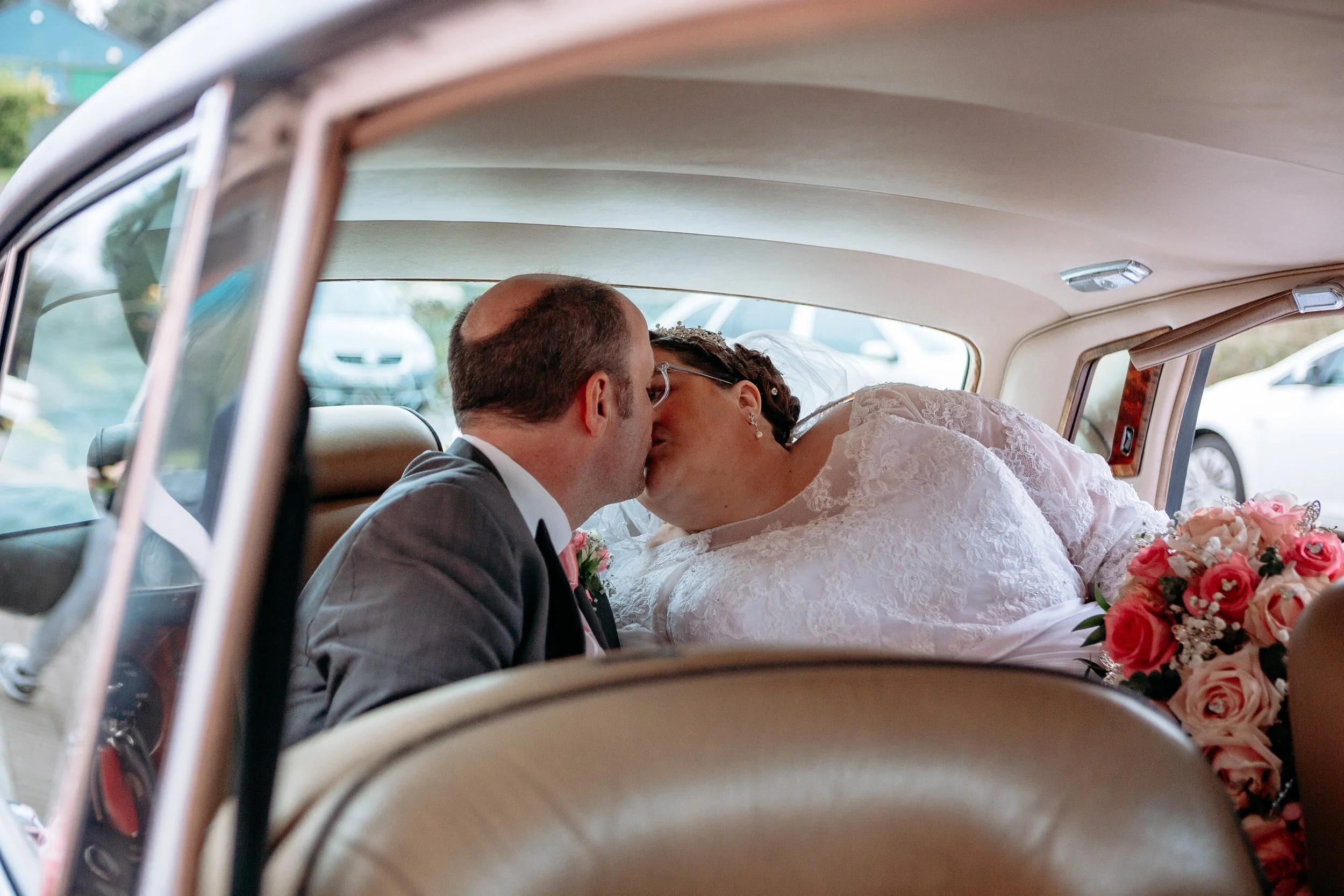 A bride and groom share a kiss inside a vintage car, with the bride holding a bouquet of pink and white roses.