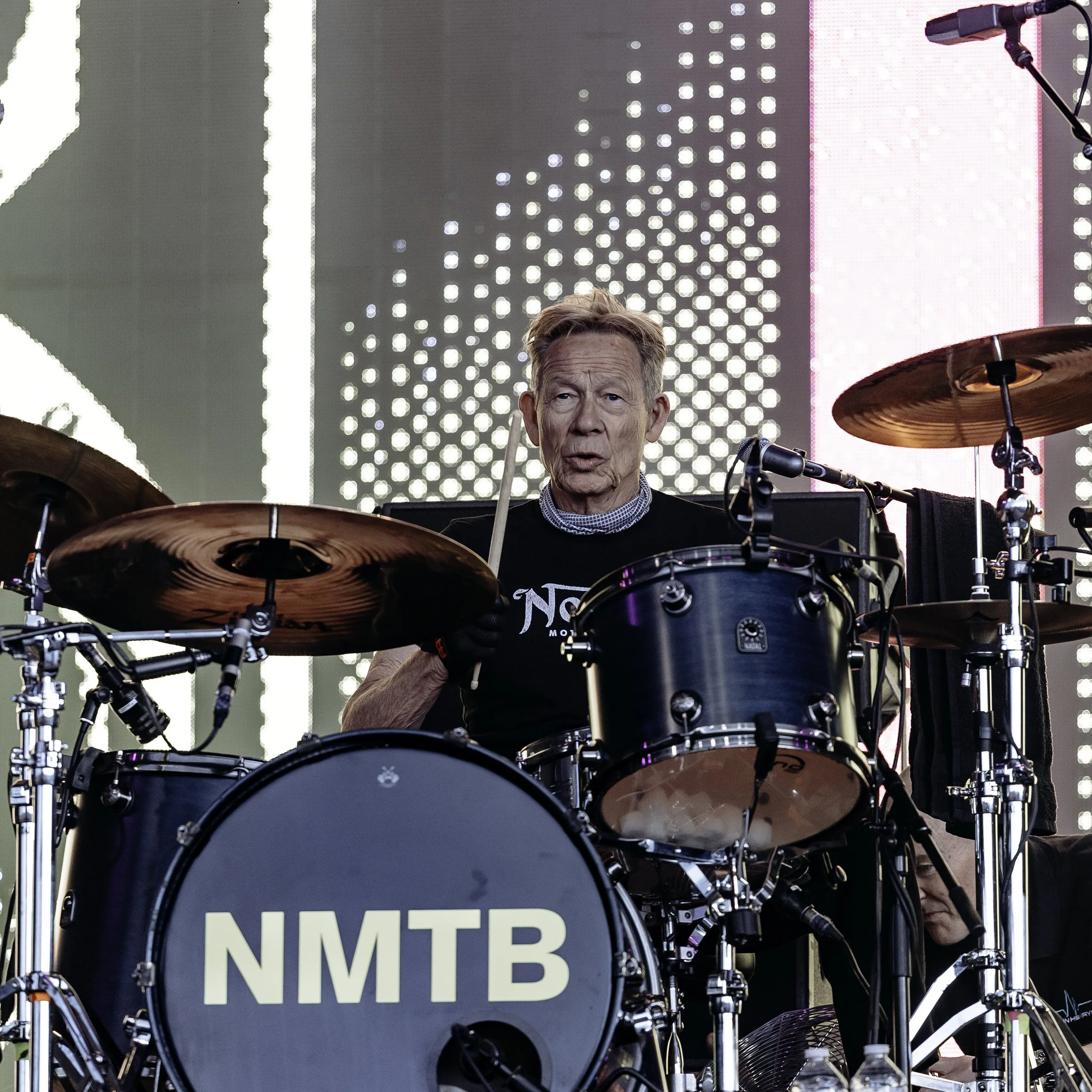 A man playing a drum set on stage with a large LED screen behind him displaying abstract lights. The drum set has the initials 'NMTB' on the bass drum.