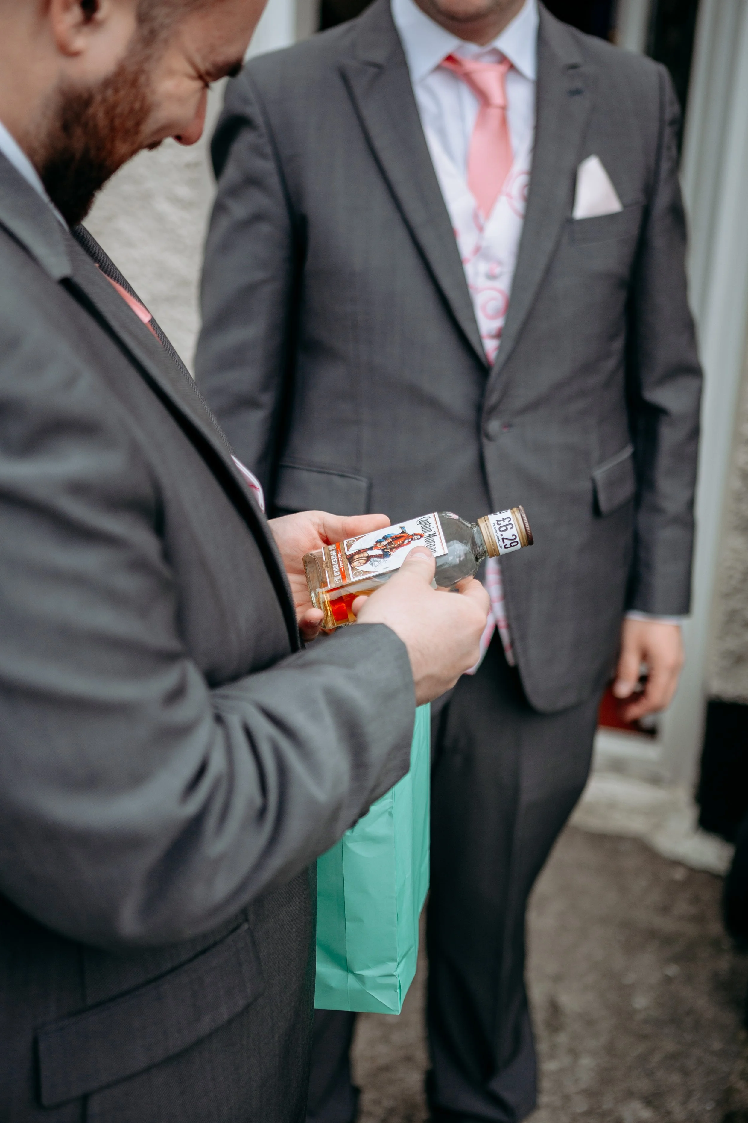 Two men dressed in suits at a social gathering, one of whom is holding a small bottle of alcoholic beverage.