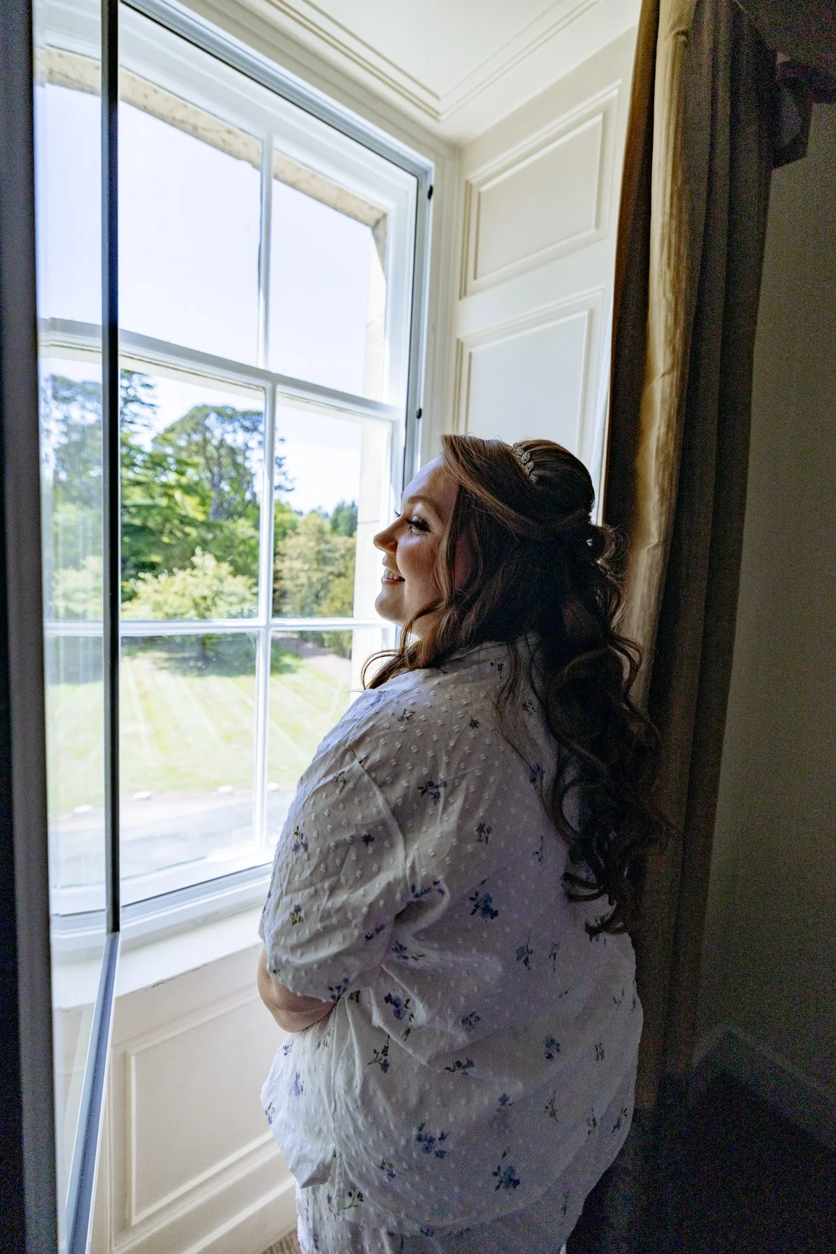 A woman with long brown hair and a headband gazing out a large window, smiling. She is wearing a white robe with small floral patterns. The window shows a scenic view of green trees and grass outside.