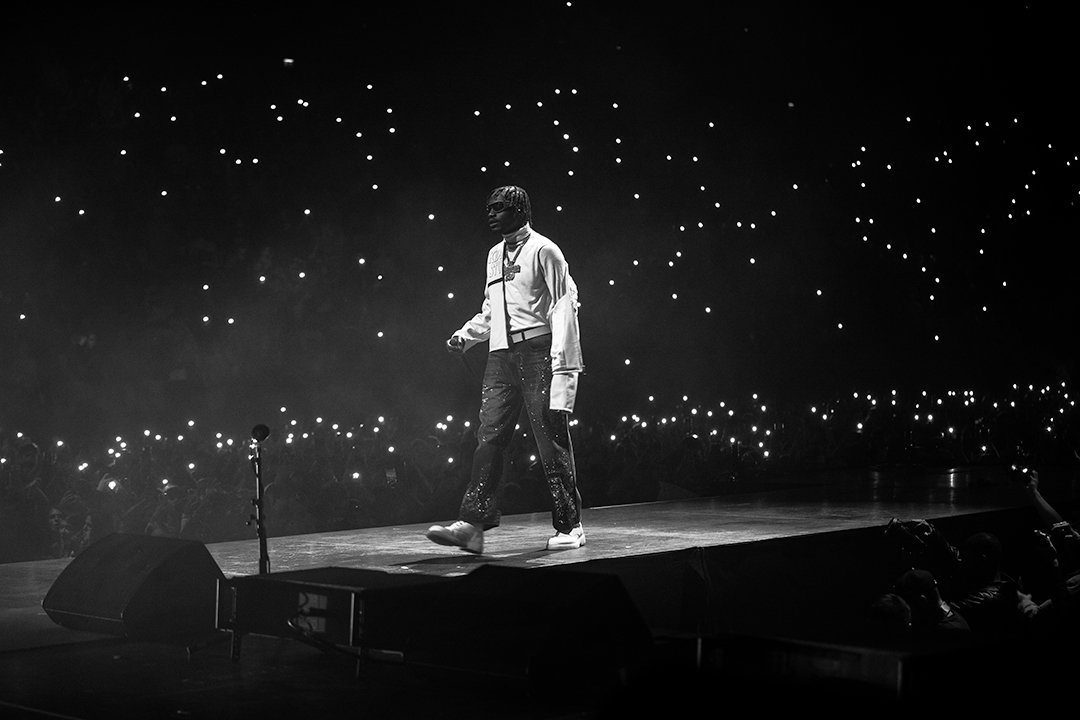 A performer walking across a stage at a concert, with many audience members holding up lights in the background, in black and white.