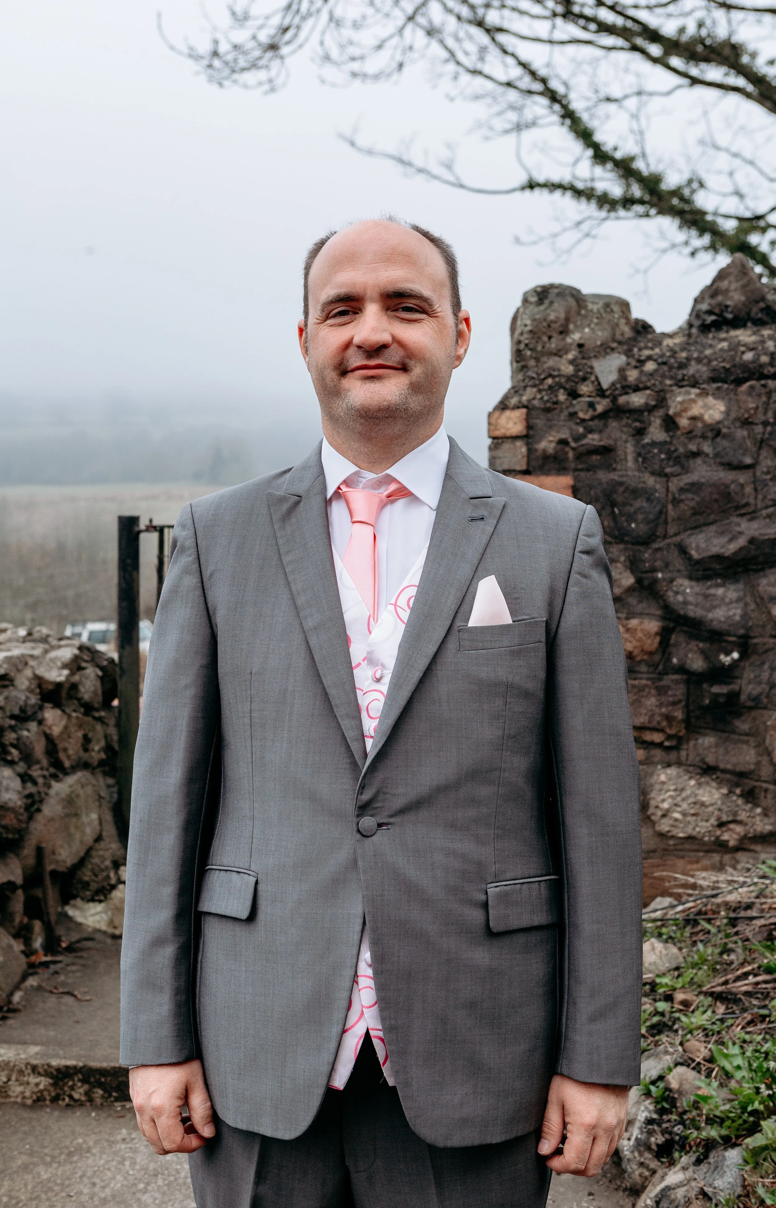 Man in gray suit with pink tie and pocket square standing outdoors in front of stone wall and foggy landscape.