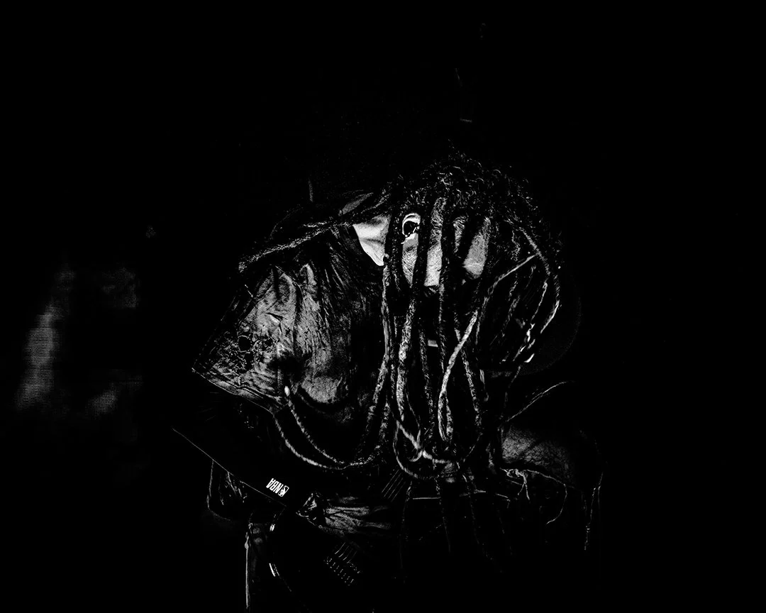 A person with long dreadlocks playing an electric guitar in a dark setting, illuminated by a single light source.