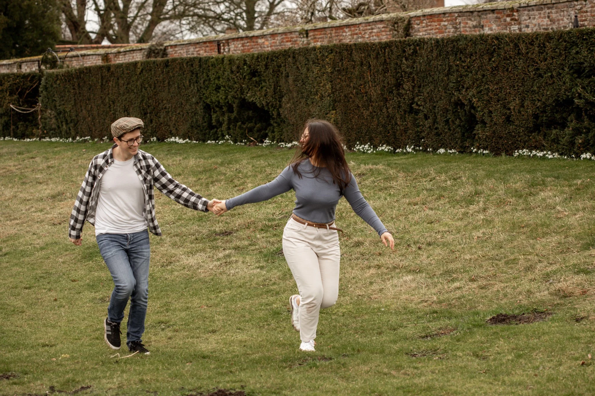 Two women holding hands and joyfully dancing or playing on a grassy field with a brick wall and bushes in the background.