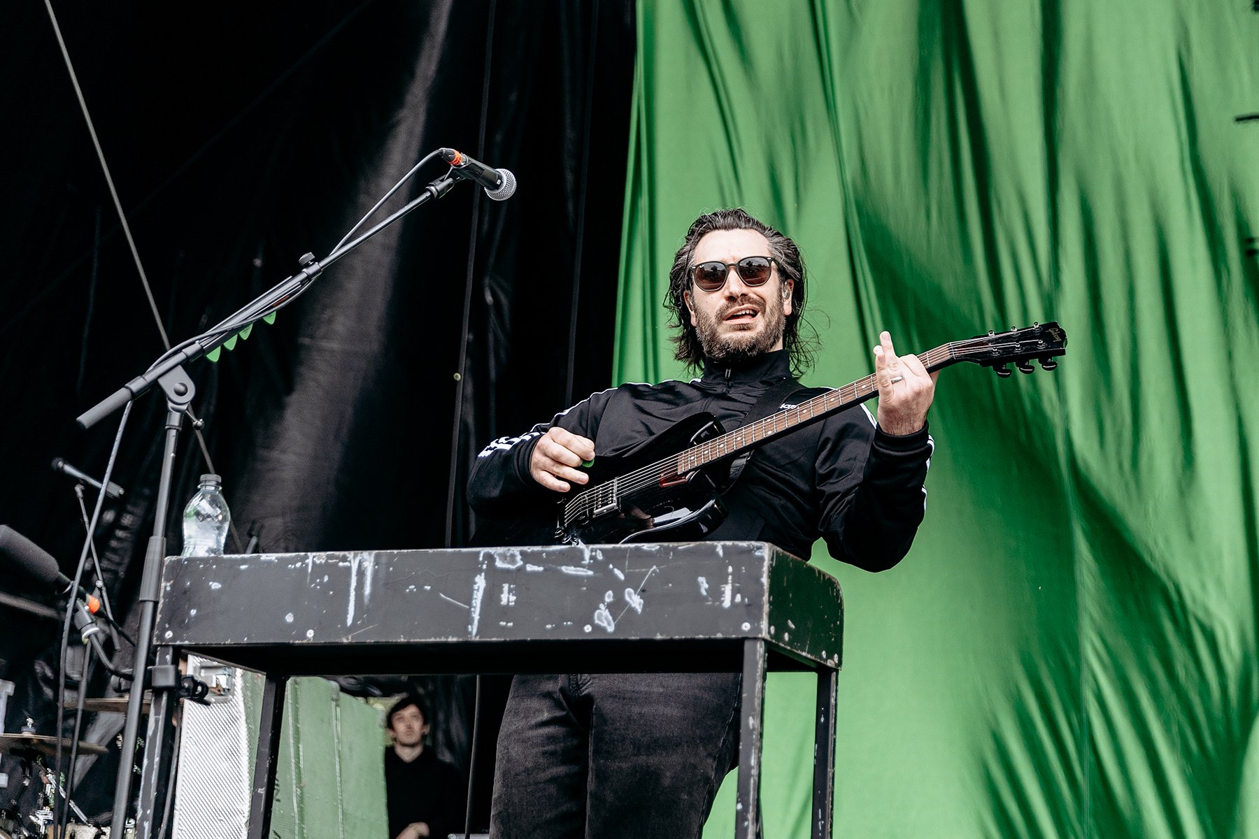 Musician with long hair and sunglasses playing an electric guitar behind a keyboard on stage with a green backdrop.