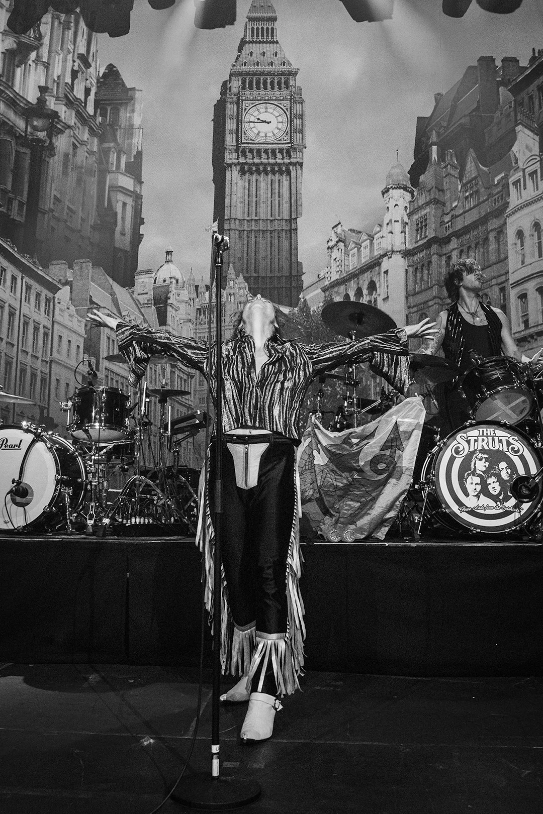 Black and white image of a band performing on stage with a backdrop of London architecture, including Big Ben. The lead singer is in focus, wearing fringed clothing, arms outstretched, and head tilted back. Drum set with "The Struts" logo visible.