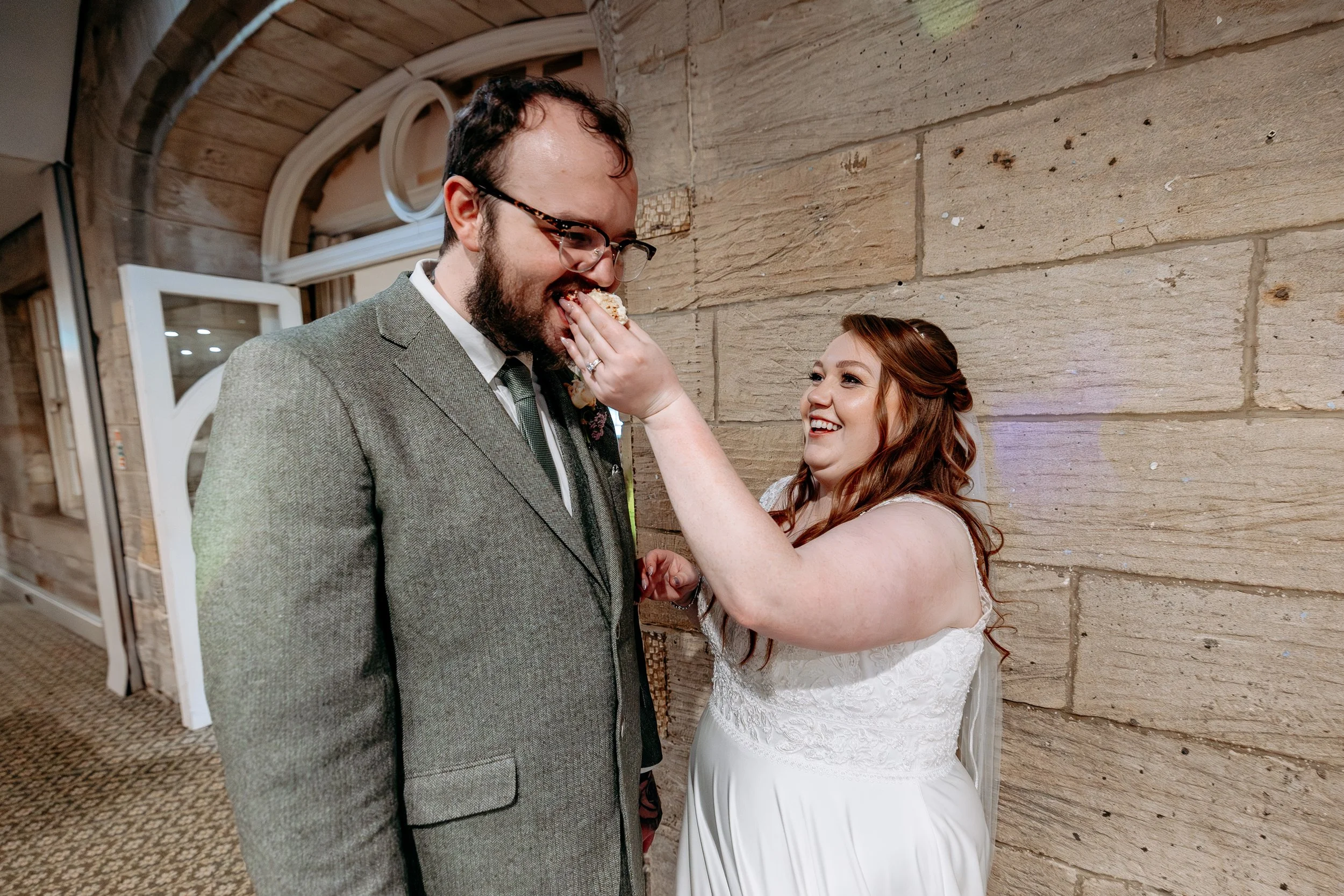 A bride feeds cake to a groom at a wedding reception, both smiling and dressed in wedding attire.