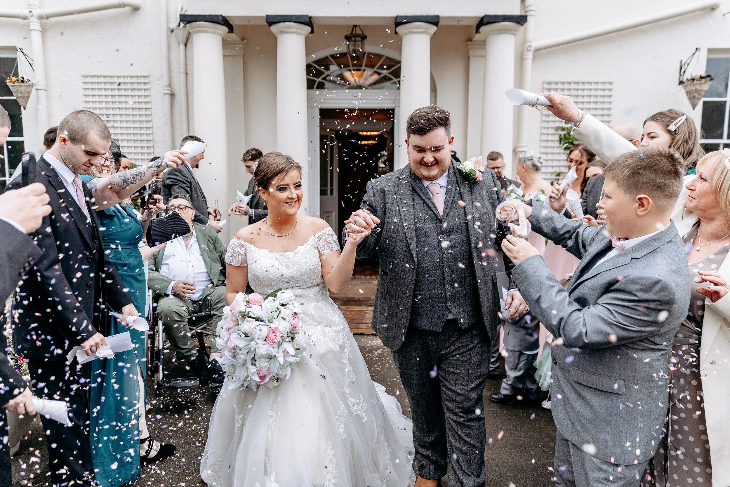A bride and groom walking out of a wedding venue, surrounded by guests throwing confetti, with the bride holding a large bouquet of white and pink flowers, and everyone celebrating.